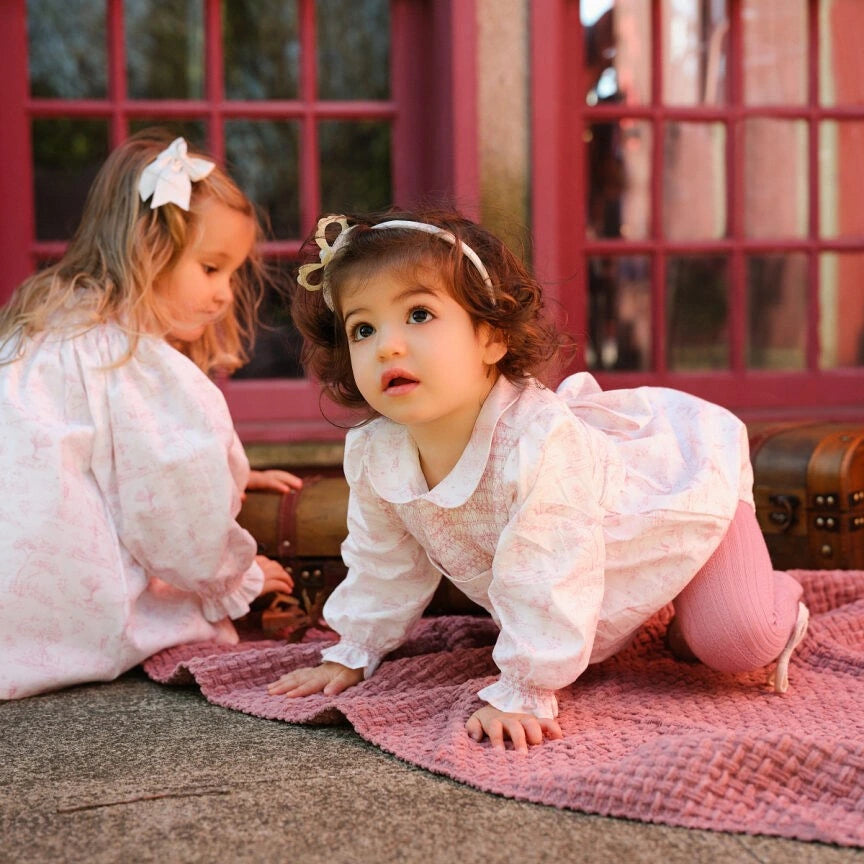 Two young girls in white dresses playing on a pink blanket in front of a red door.
