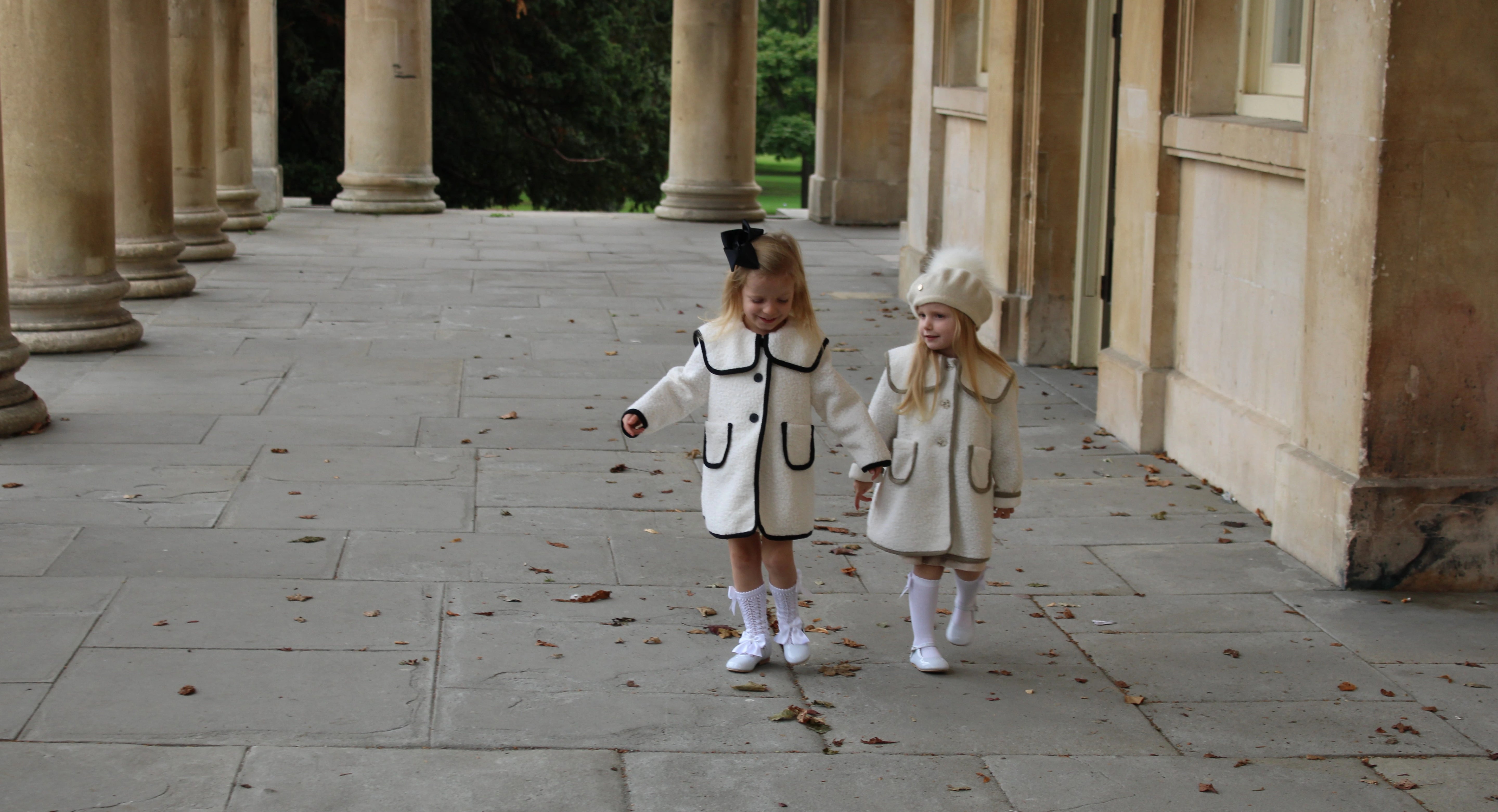 Two children in matching outfits walking on a stone path with columns in the background.