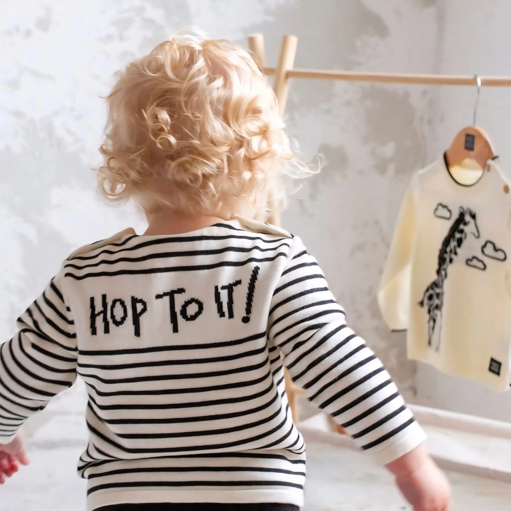 Child wearing a striped shirt with 'Hop to it!' text, standing in front of a clothing rack with baby clothes.