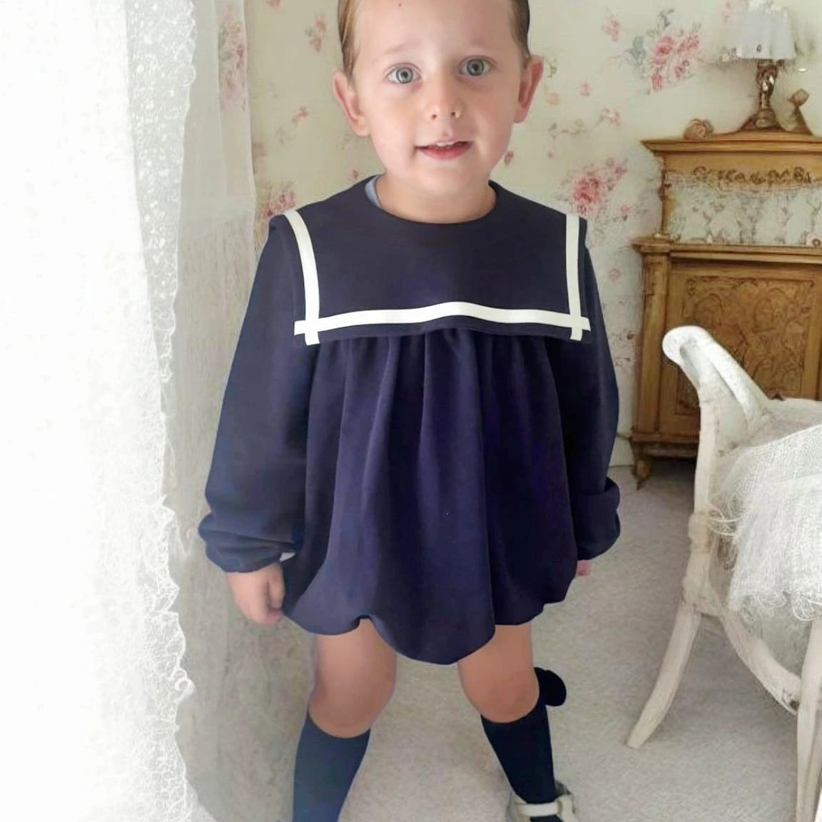 Child wearing a navy dress with white trim in a room with floral wallpaper.