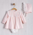 Pink baby outfit with top and pants hanging on a wooden hanger against a light wooden background.