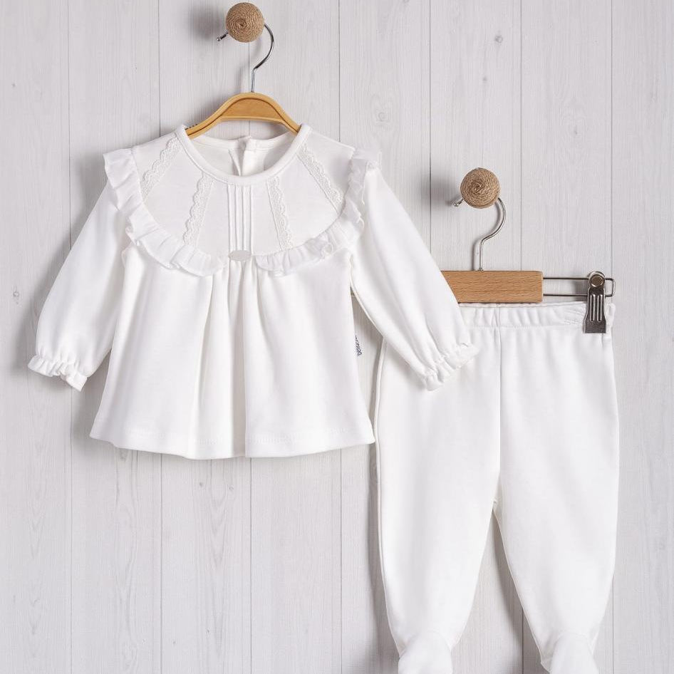 White children's outfit with a top and pants on wooden hangers against a light wooden panel background.