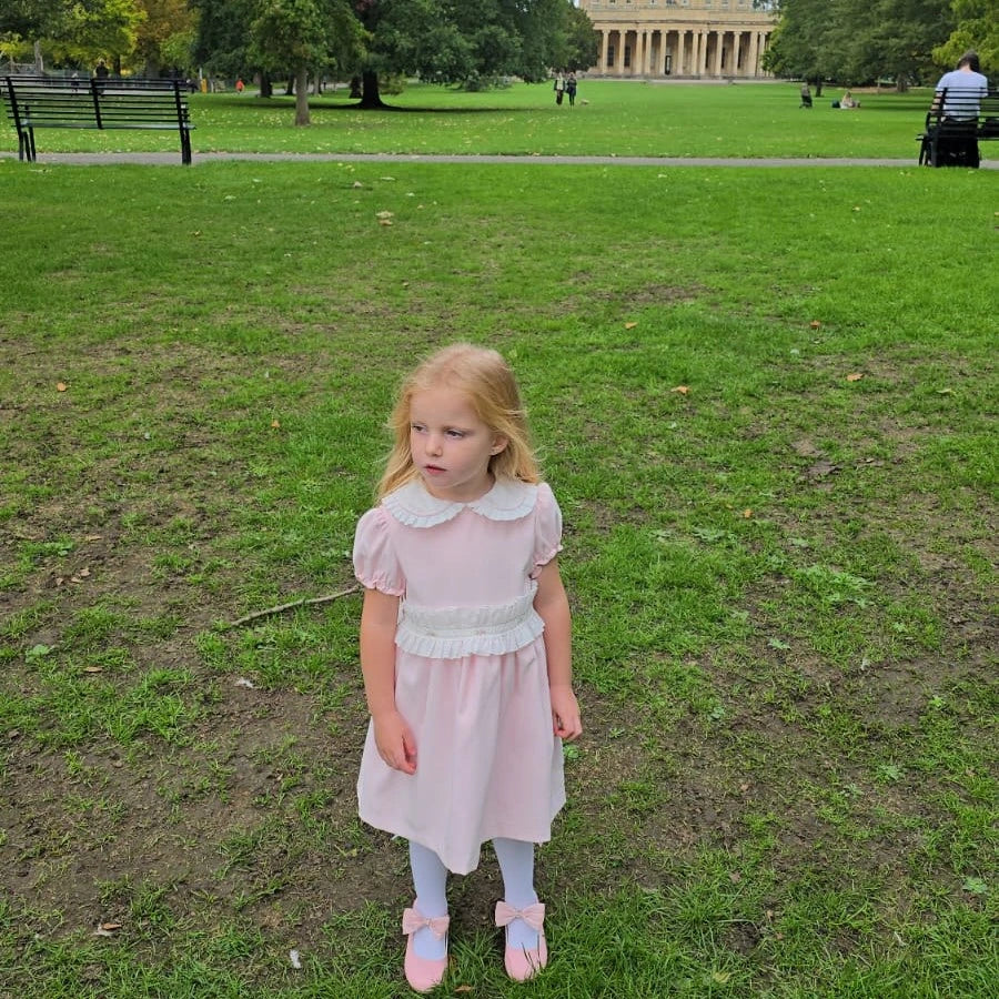 Young girl in a pink dress standing on a grassy field with trees and a building in the background.