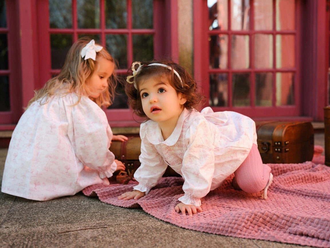 Two young girls in white dresses playing on a pink blanket in front of a red door.