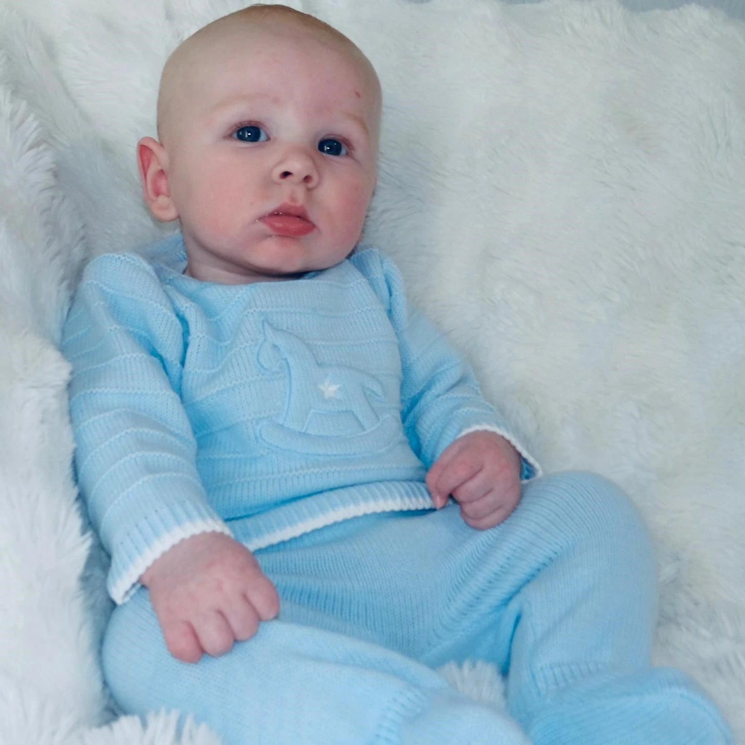 Baby wearing a light blue knitted outfit on a white fluffy blanket