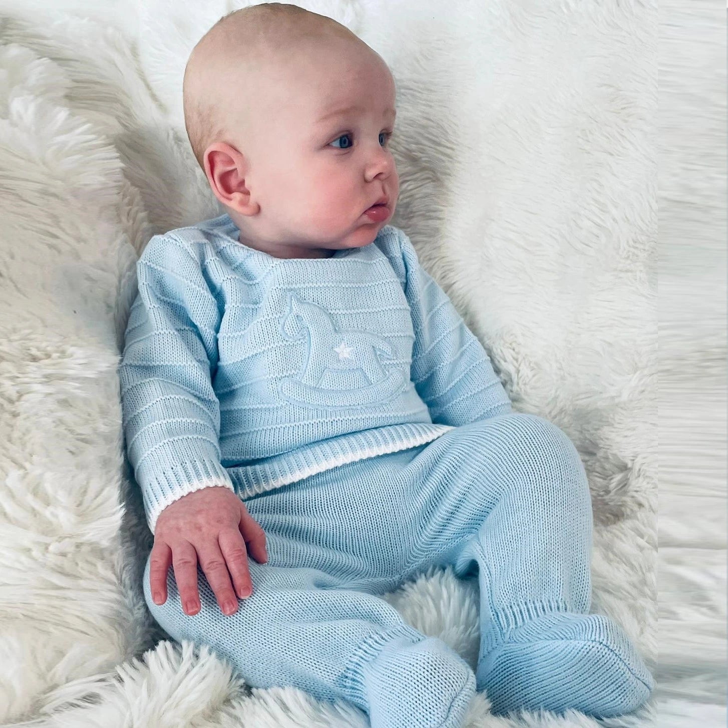 Baby in a light blue knitted outfit lying on a white fluffy surface