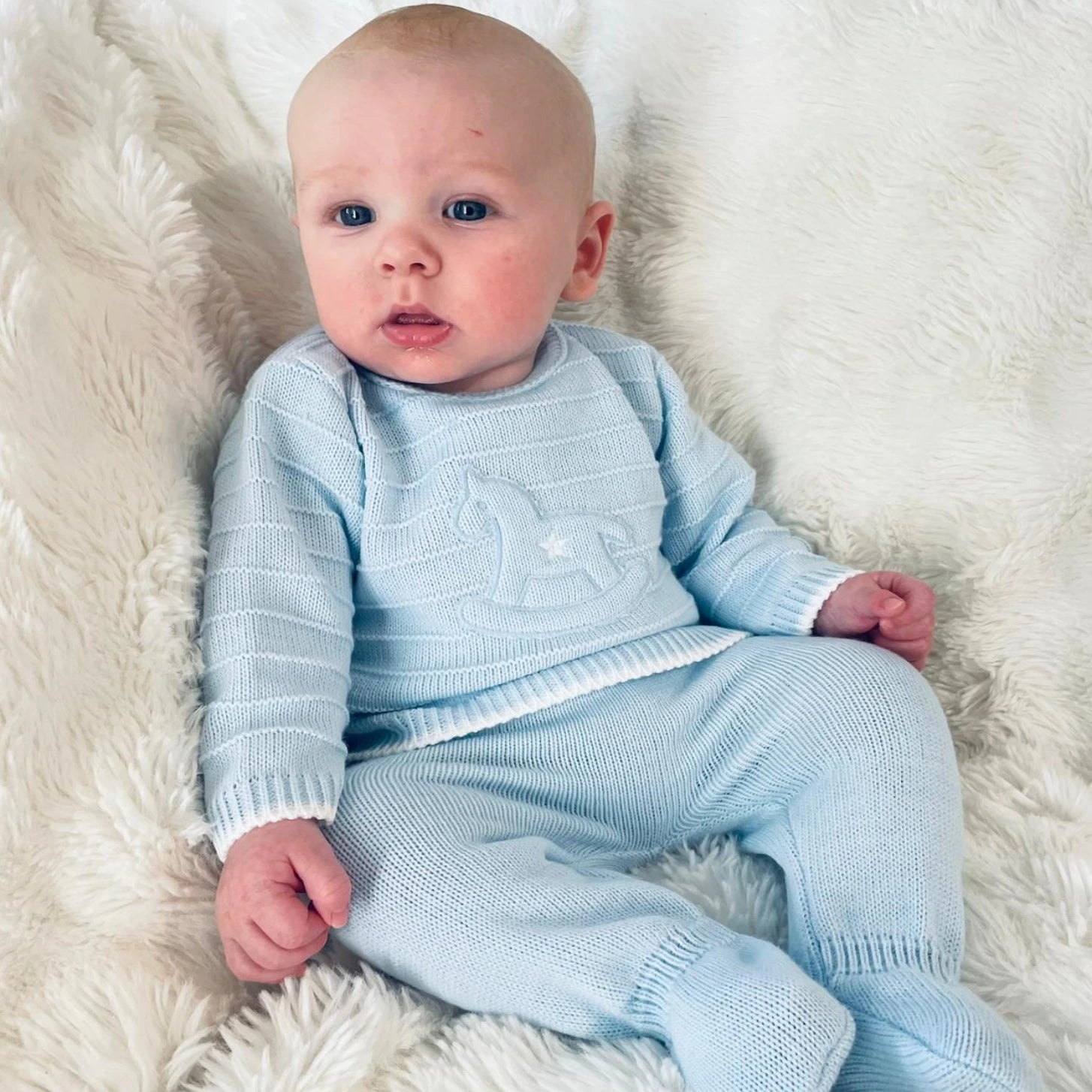 Baby in a light blue knitted outfit lying on a white fluffy surface