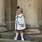 Young girl in a white dress with black trim standing in front of stone columns.