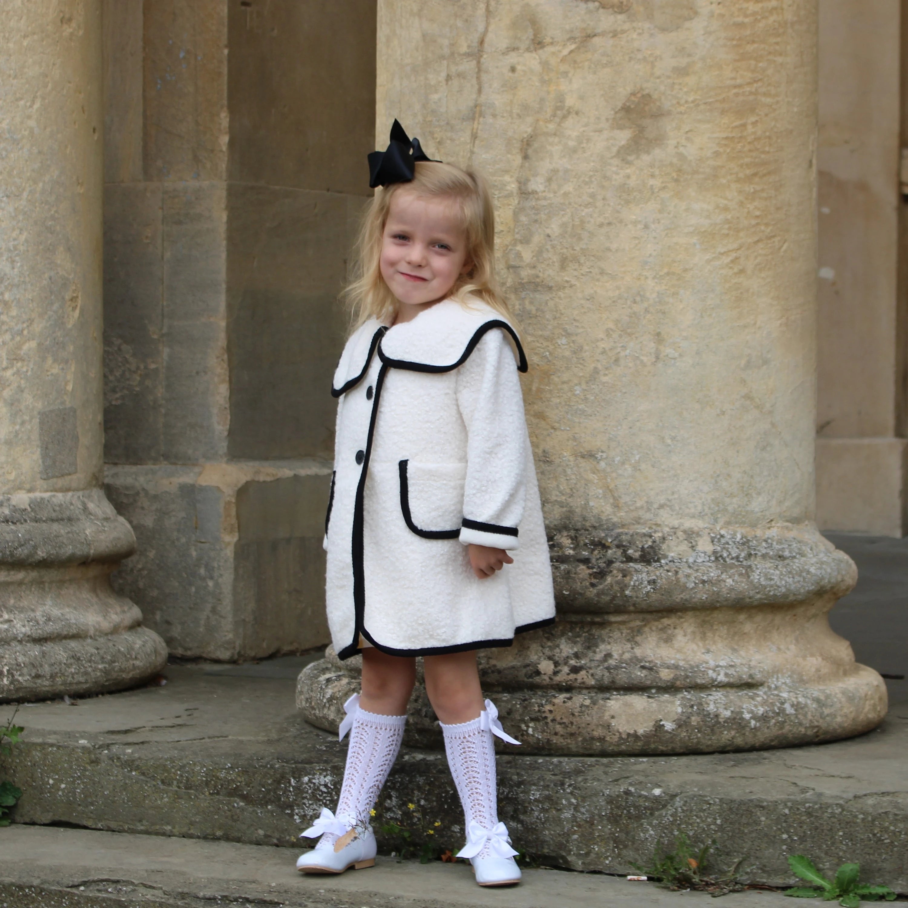 Young girl in a white dress with black trim standing in front of stone columns.