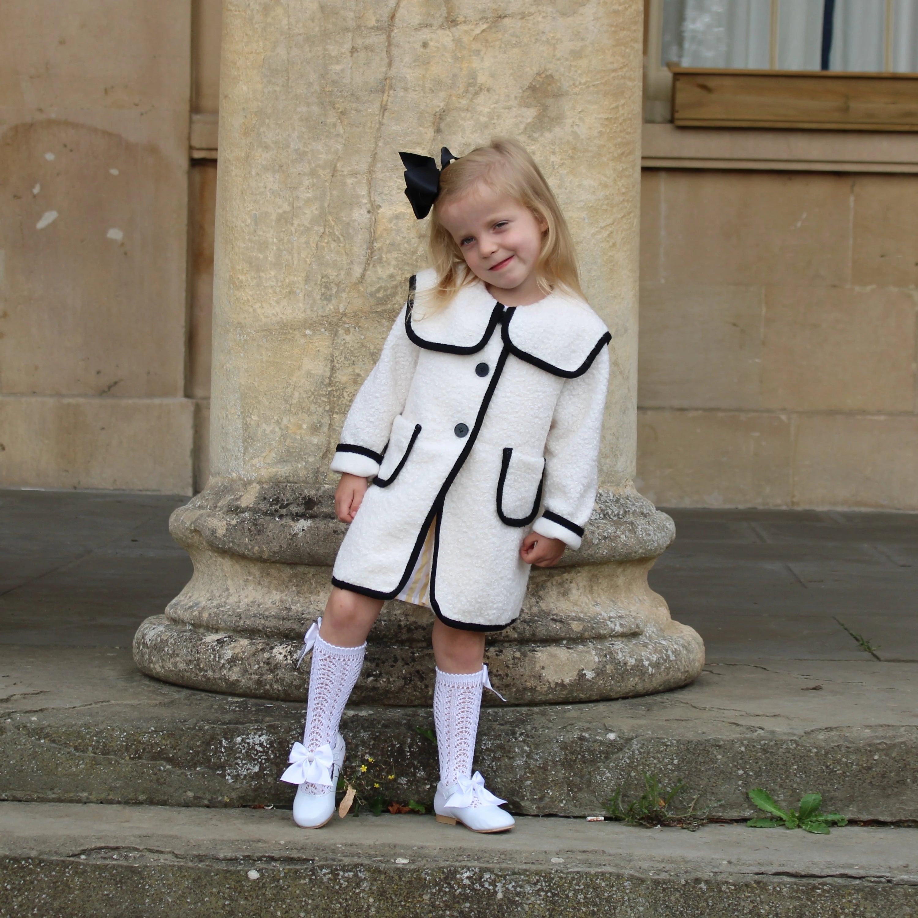Young girl in a white coat with black trim standing on stone steps.