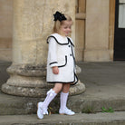 Young girl in a white dress with black trim standing outdoors near stone columns.