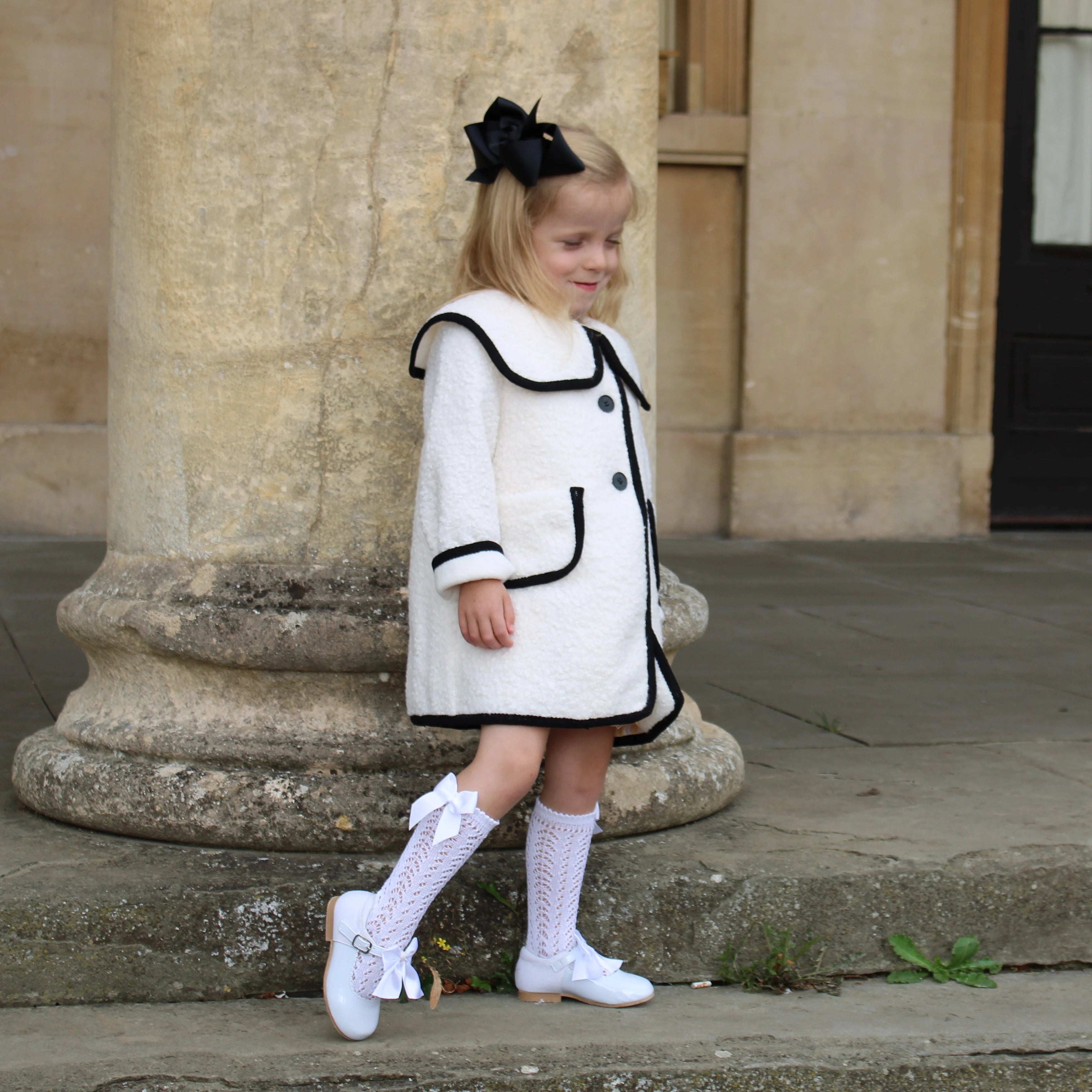Young girl in a white dress with black trim standing outdoors near stone columns.