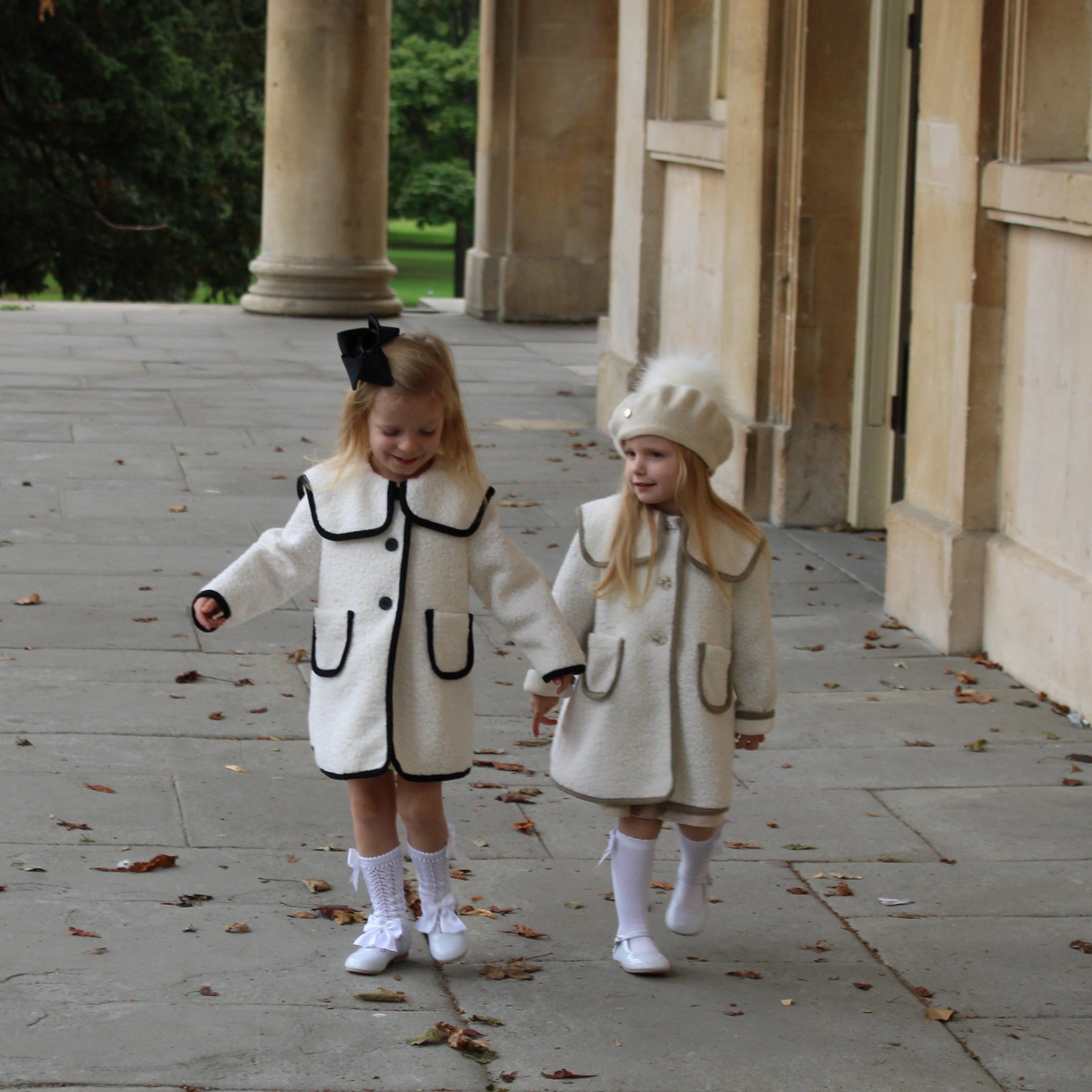 Two young girls in matching white coats walking outdoors on a stone path.
