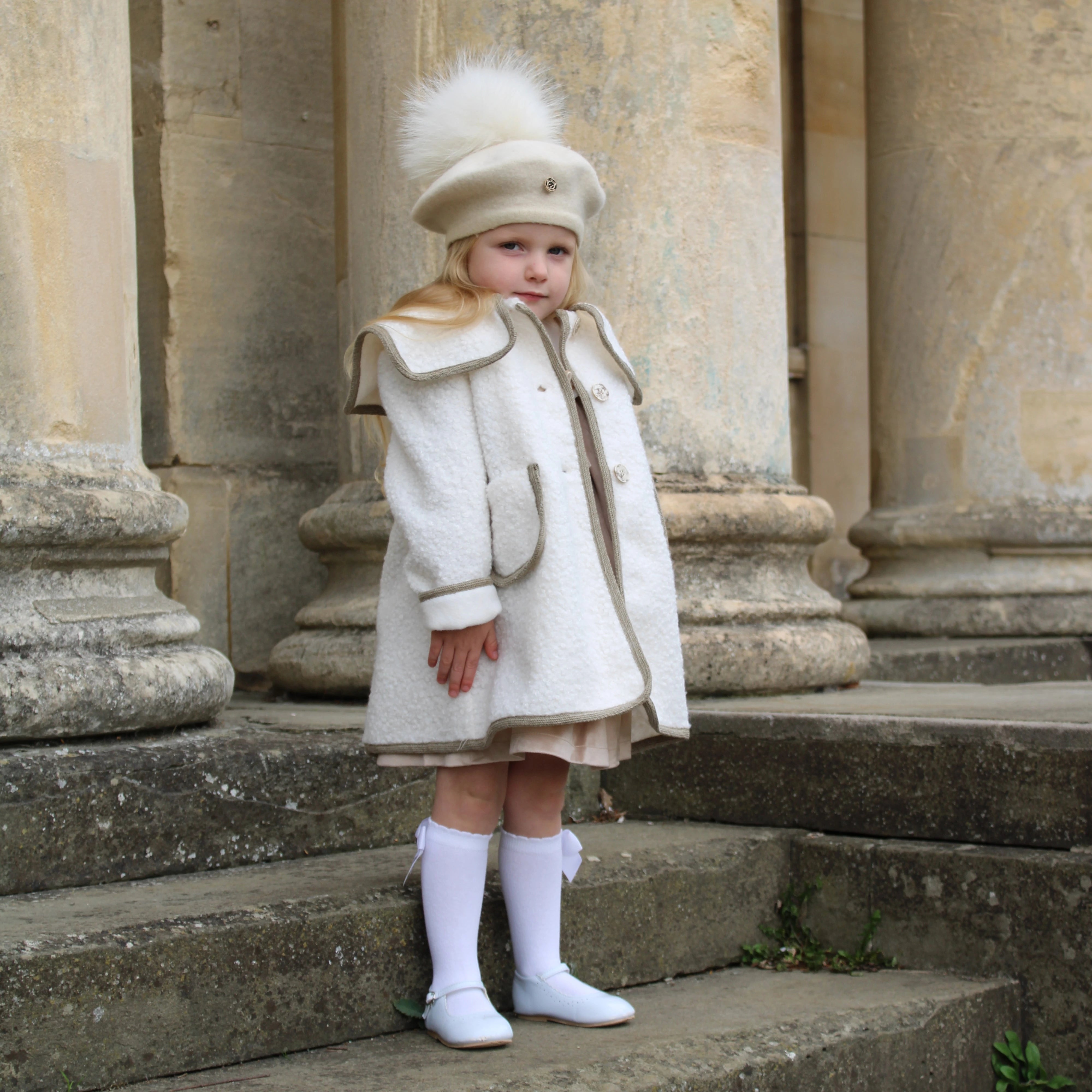 Child in a white coat and hat with a fur pom-pom standing on stone steps.