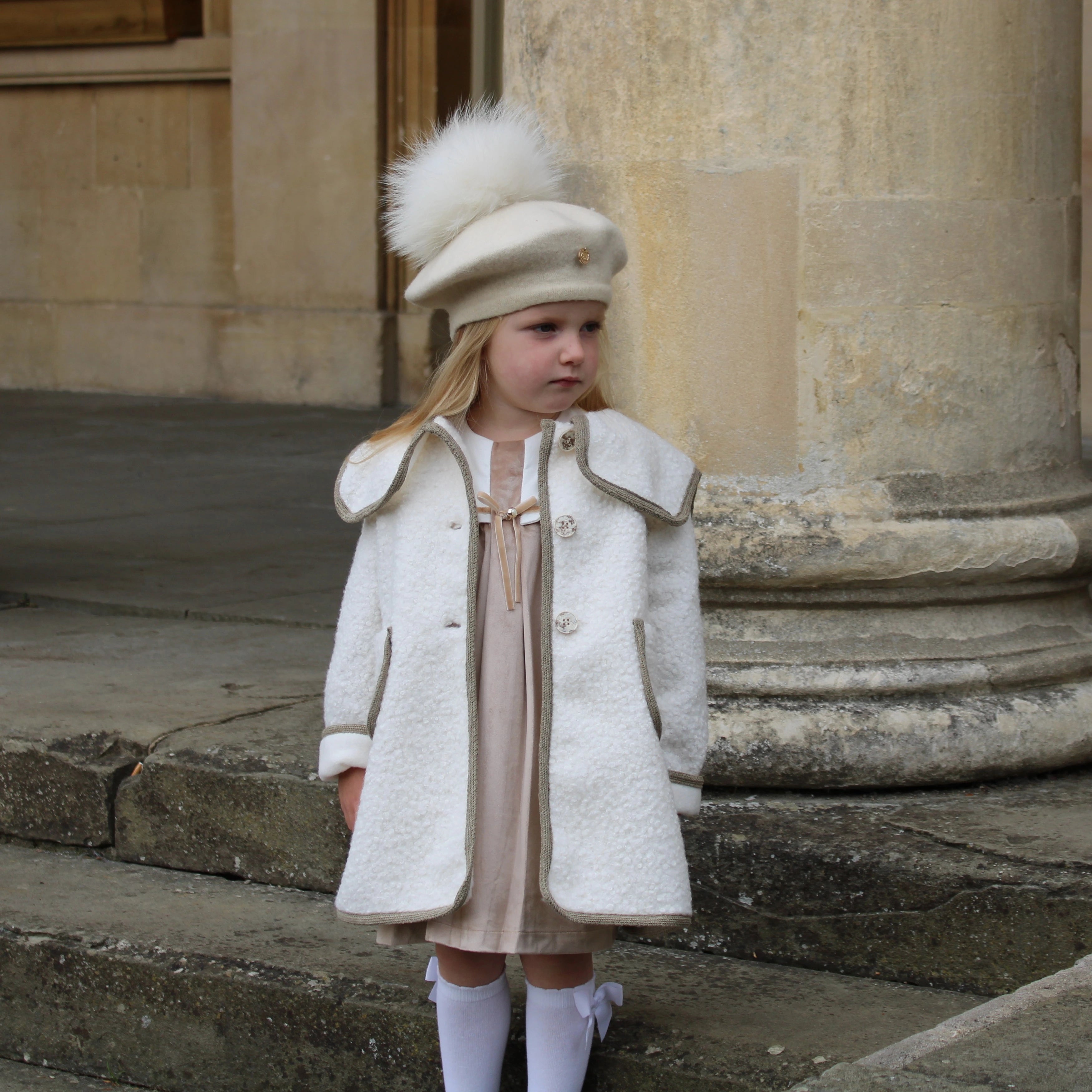 Young girl in a white coat and hat standing on stone steps.