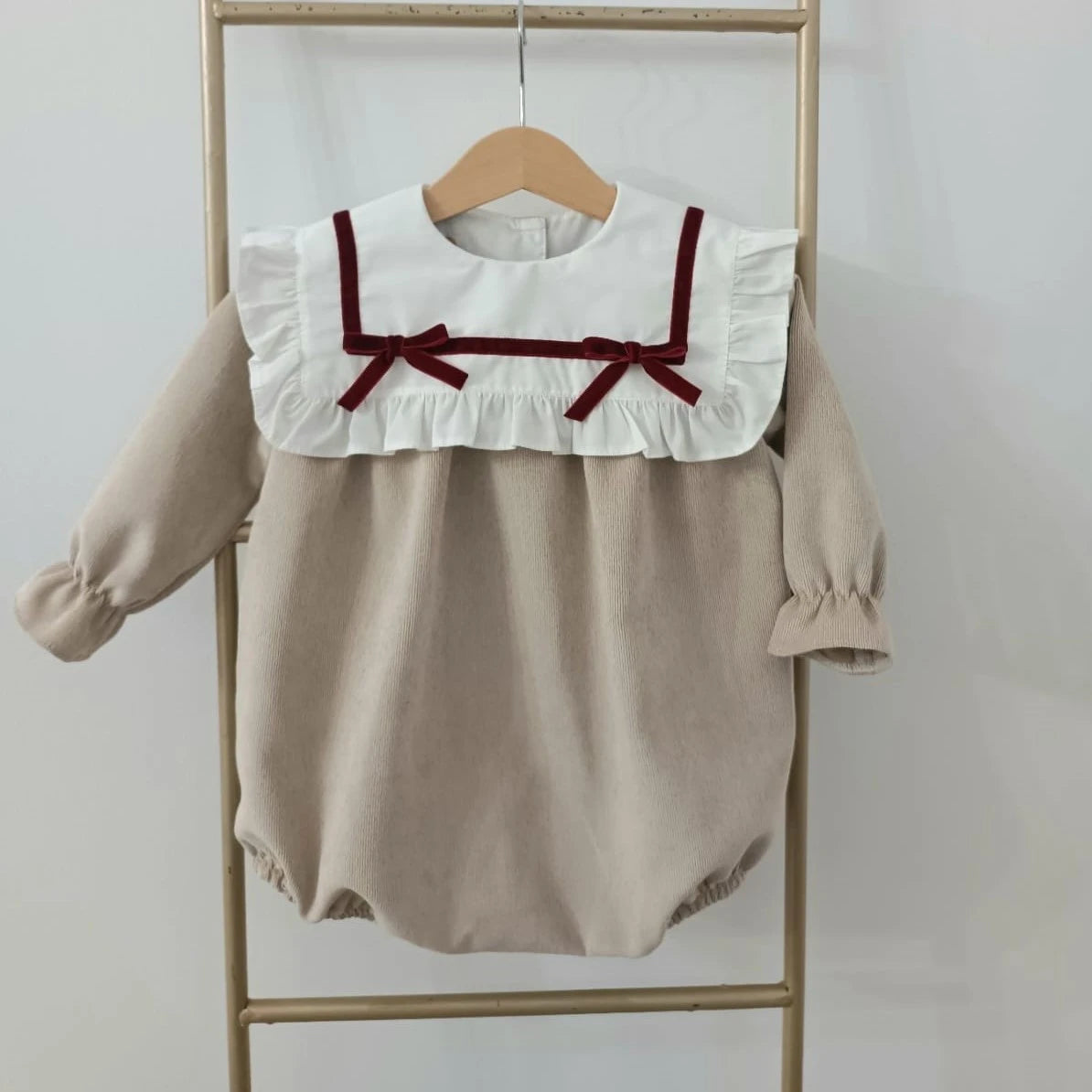 Beige baby romper with white collar and red trim hanging on a wooden rack against a white background
