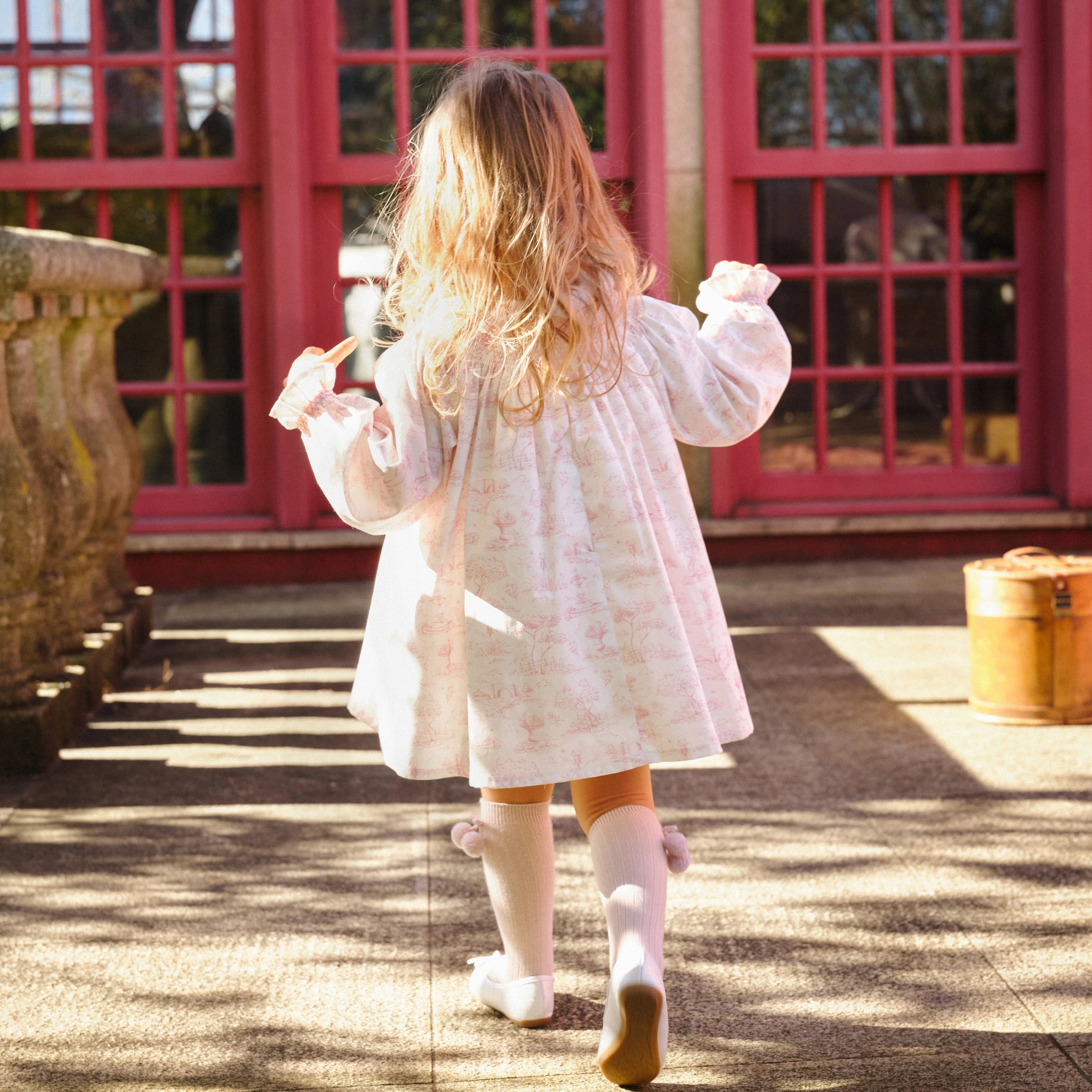 Child in a white dress walking outdoors with a pink building in the background