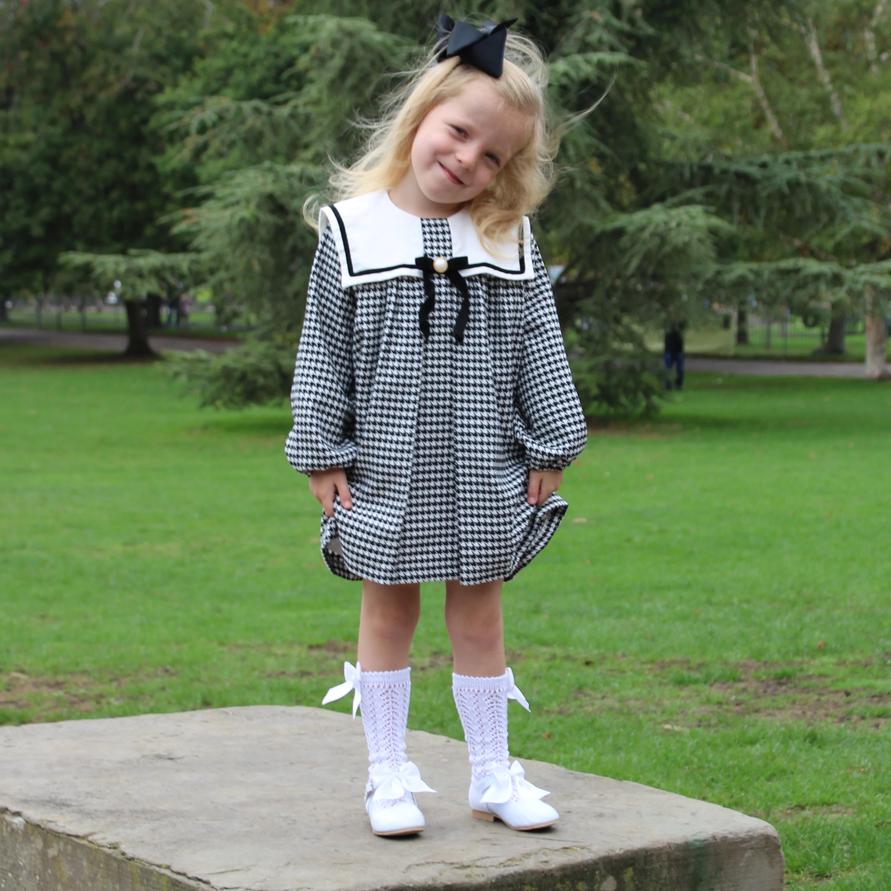 Young girl in a checkered dress with a sailor collar standing on a stone block in a park.
