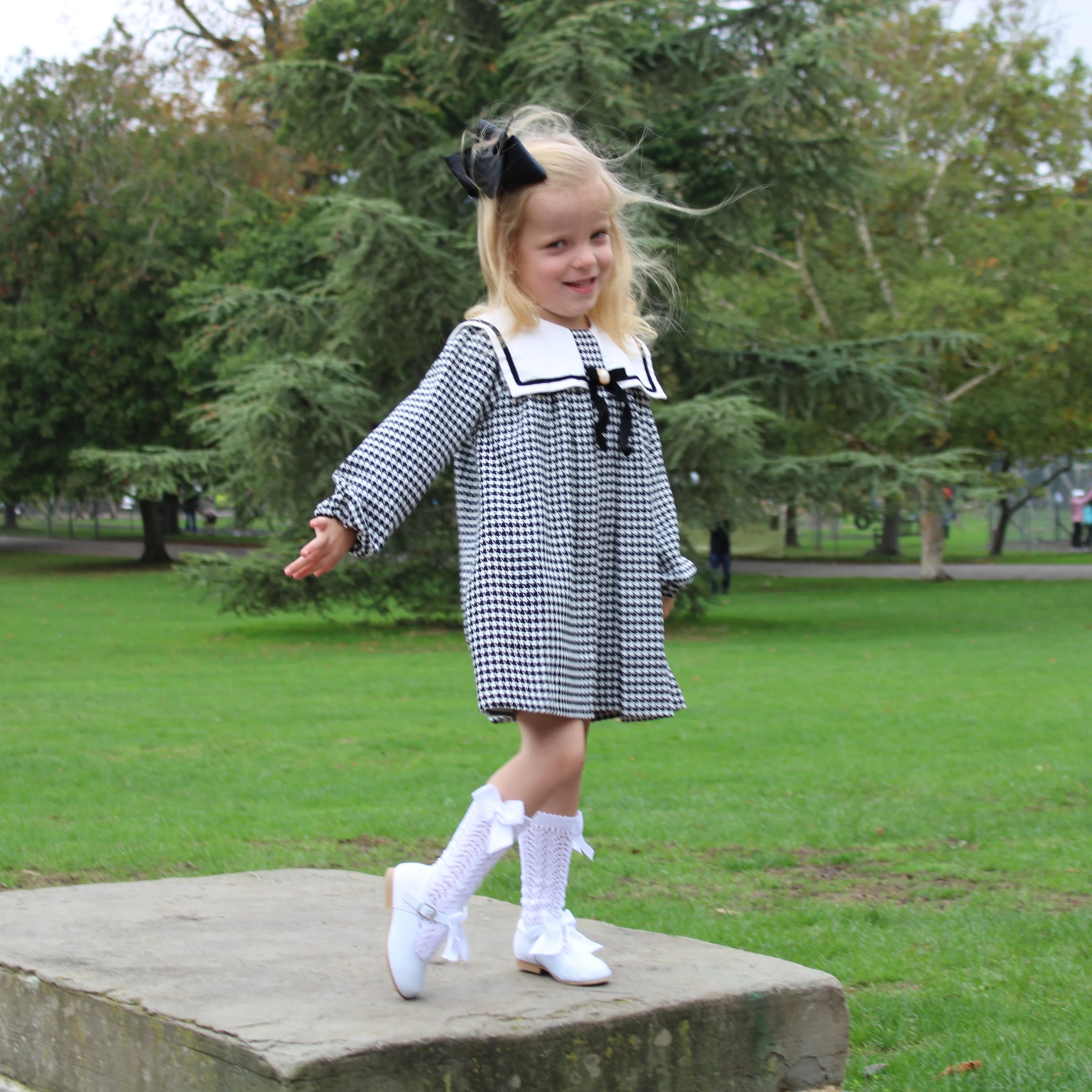 Young girl in a black and white checkered dress with a bow standing on a stone block in a park.