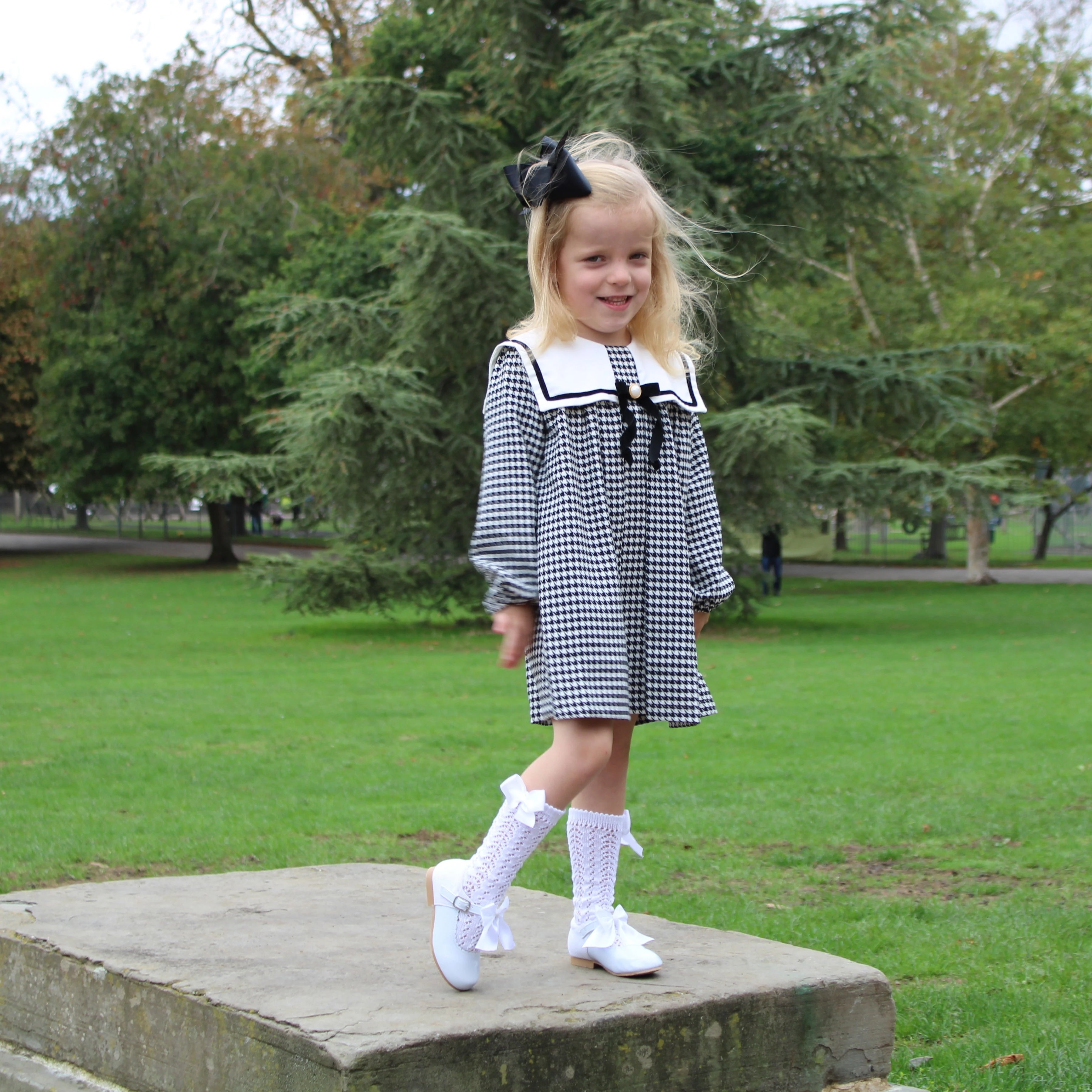 Young girl in a checkered dress with a bow standing on a stone pedestal in a park.