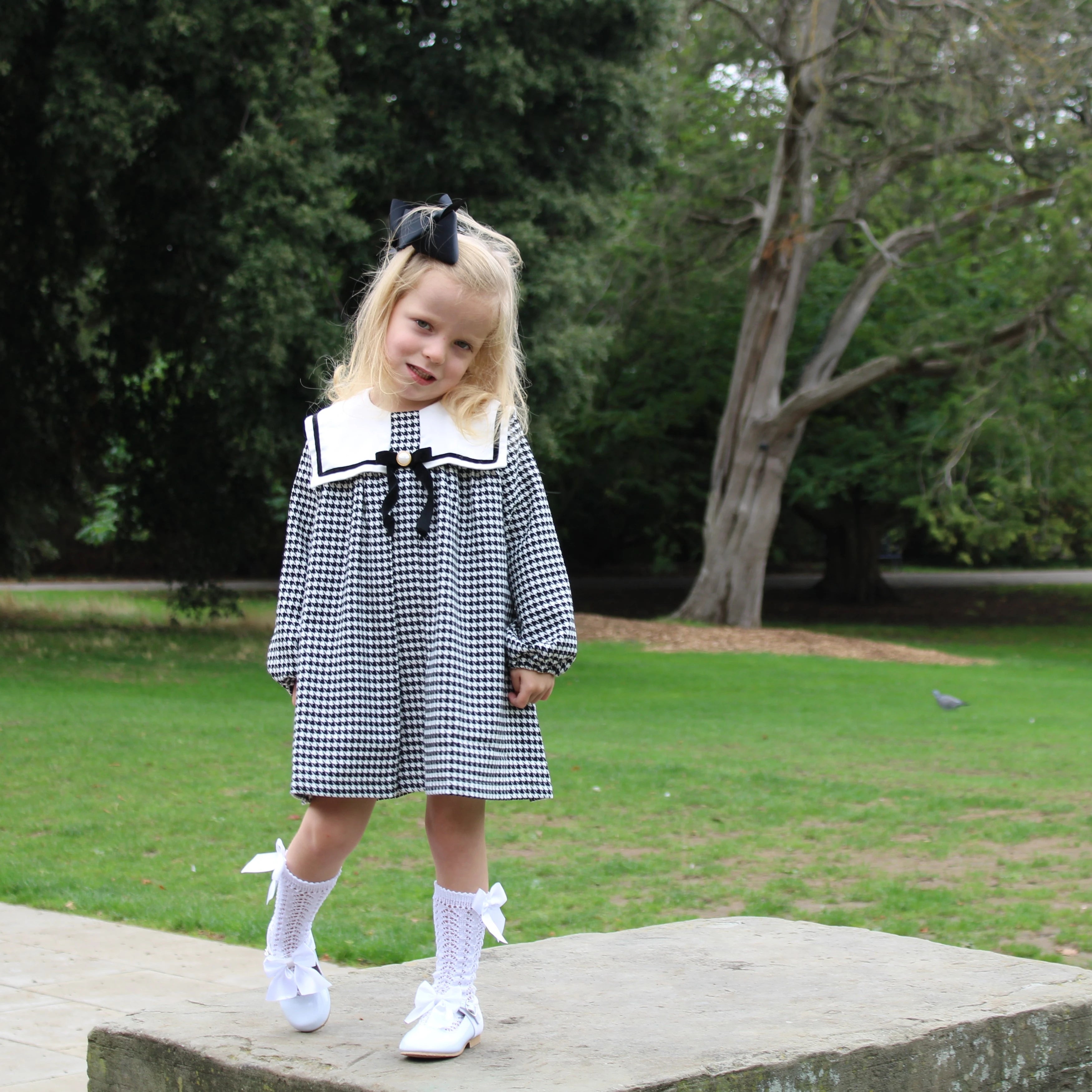 Young girl in a black and white checkered dress with a bow standing on a stone path in a park.