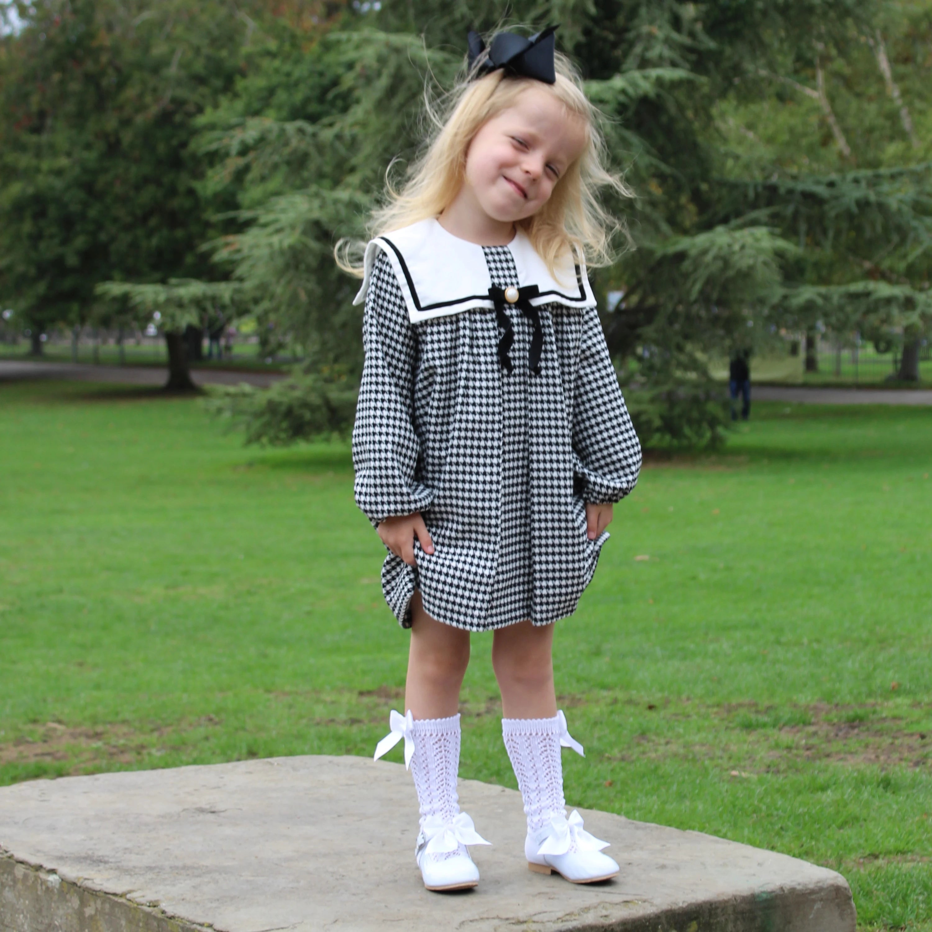 Young girl in a black and white checkered dress with a bow standing on a stone platform in a park.