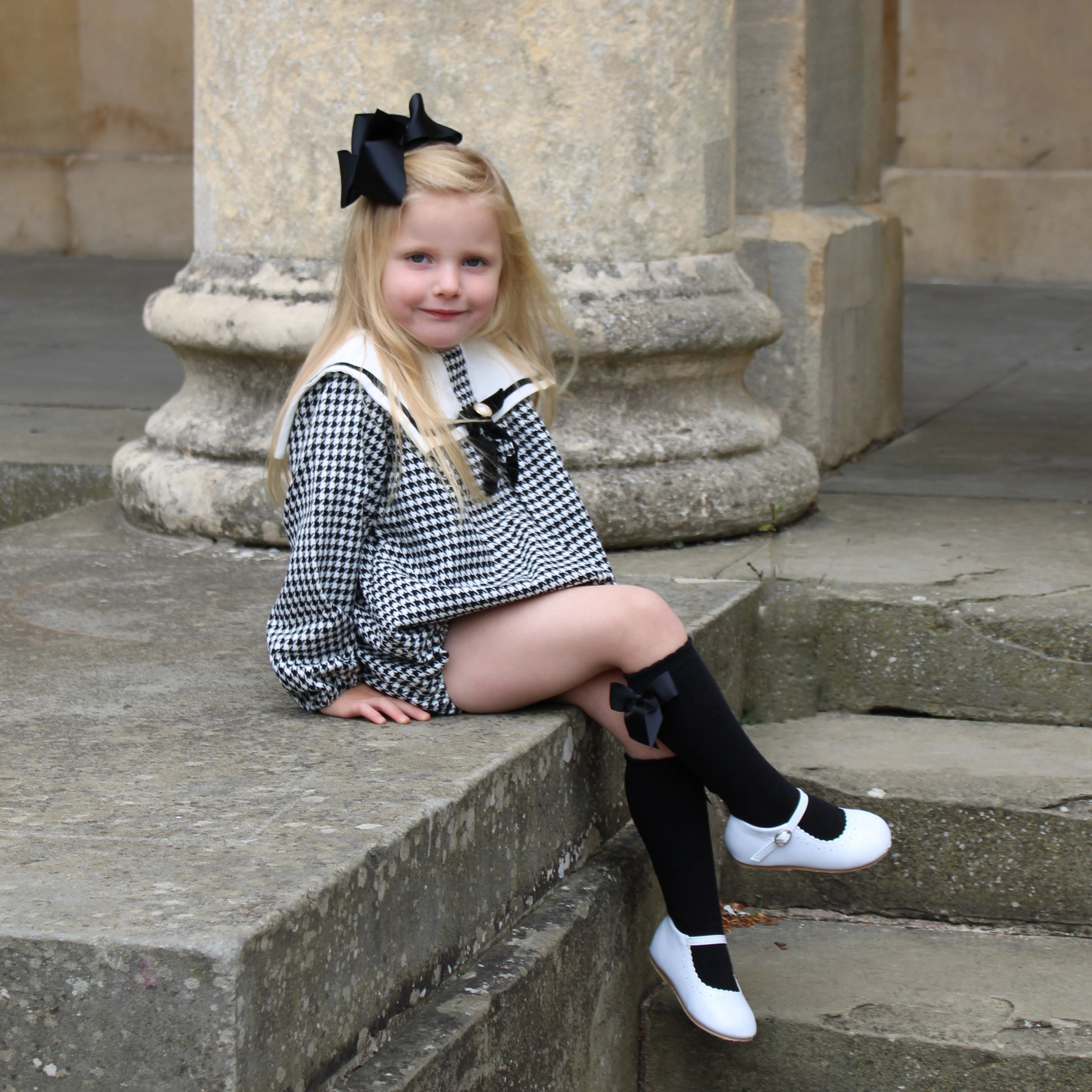 Young girl in a checkered outfit with a large bow sitting on stone steps.