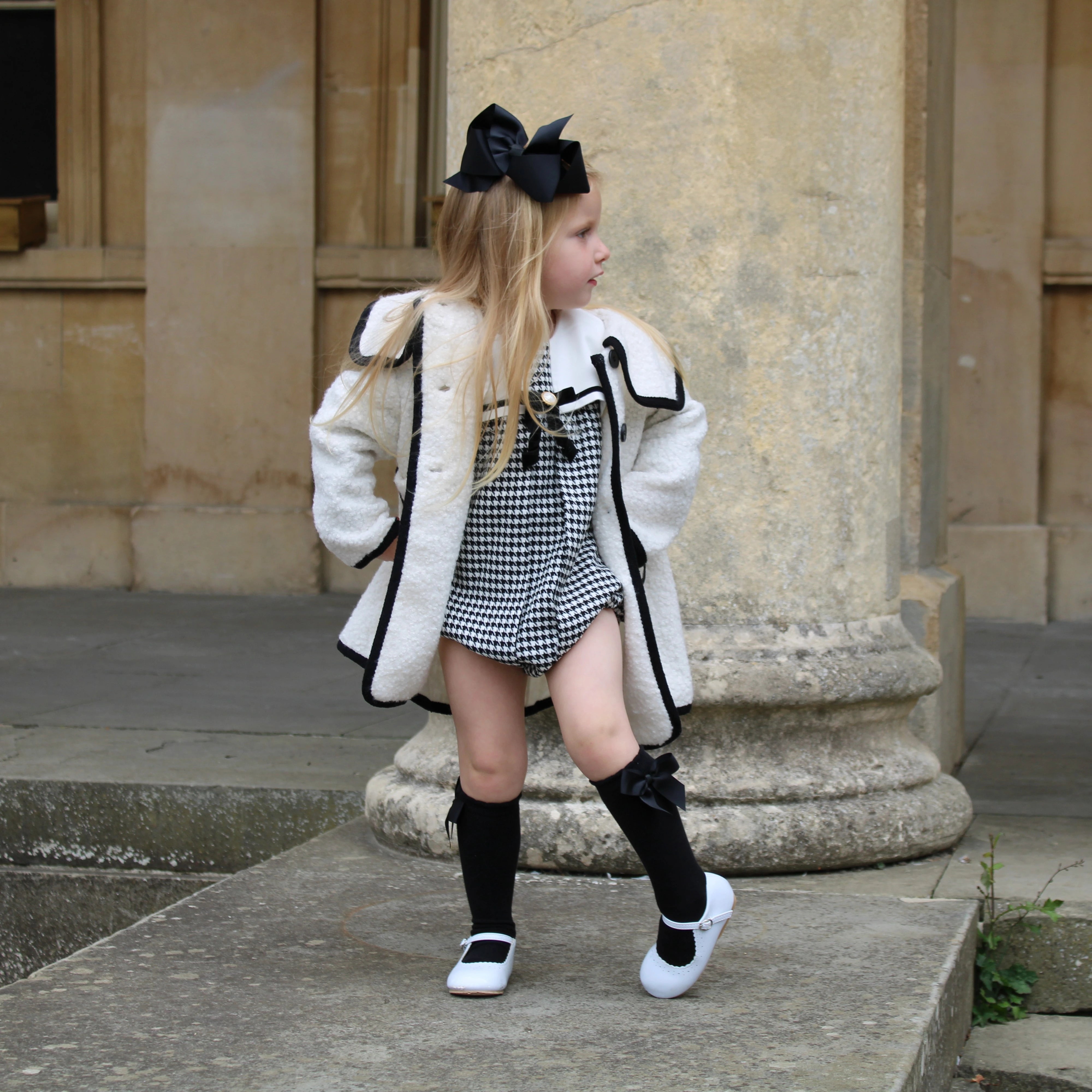 Young girl in a white coat with black trim, black knee-high socks, and white shoes standing on stone steps.