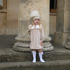 Young girl in a beige dress and white hat standing in front of stone columns.