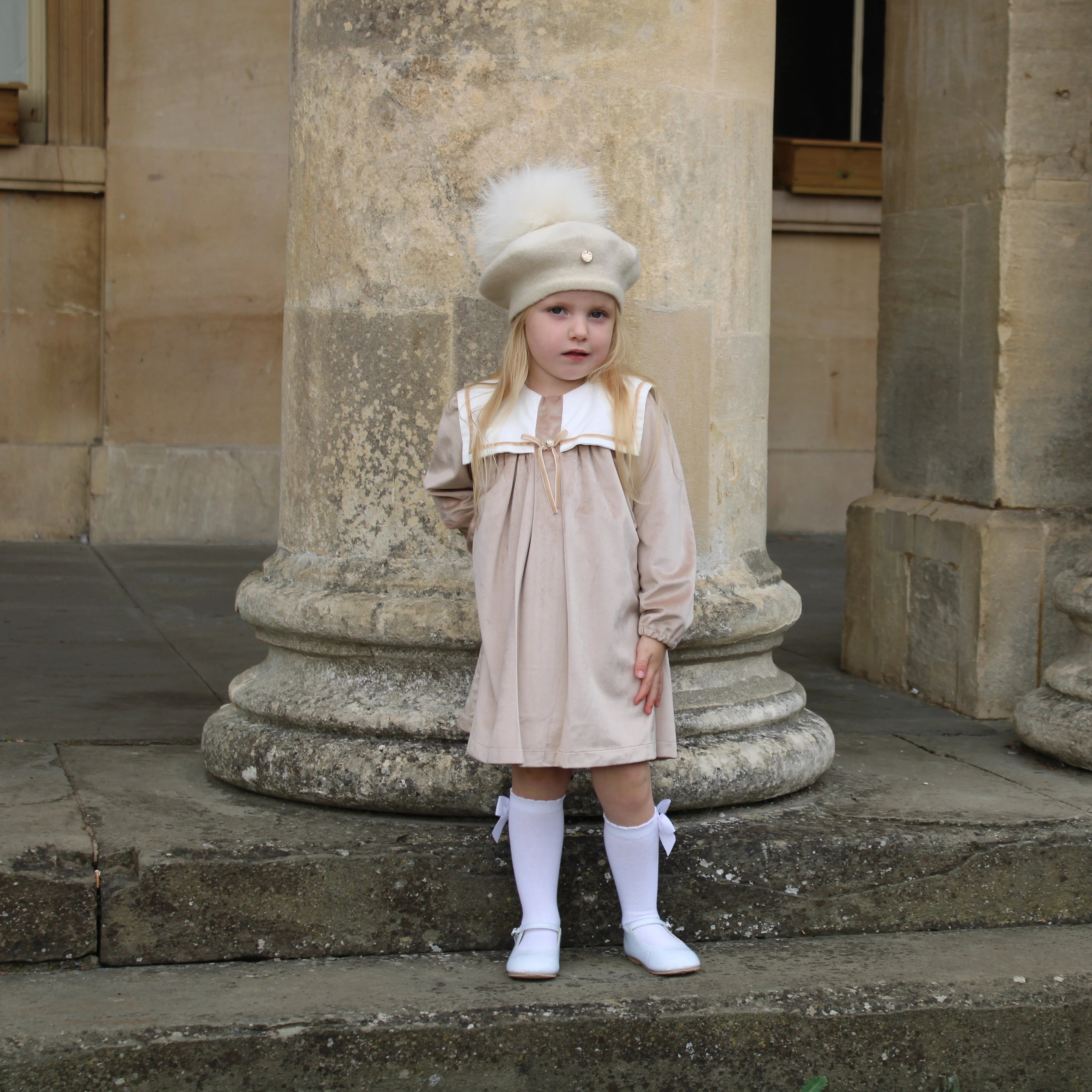 Young girl in a beige dress and white hat standing in front of stone columns.