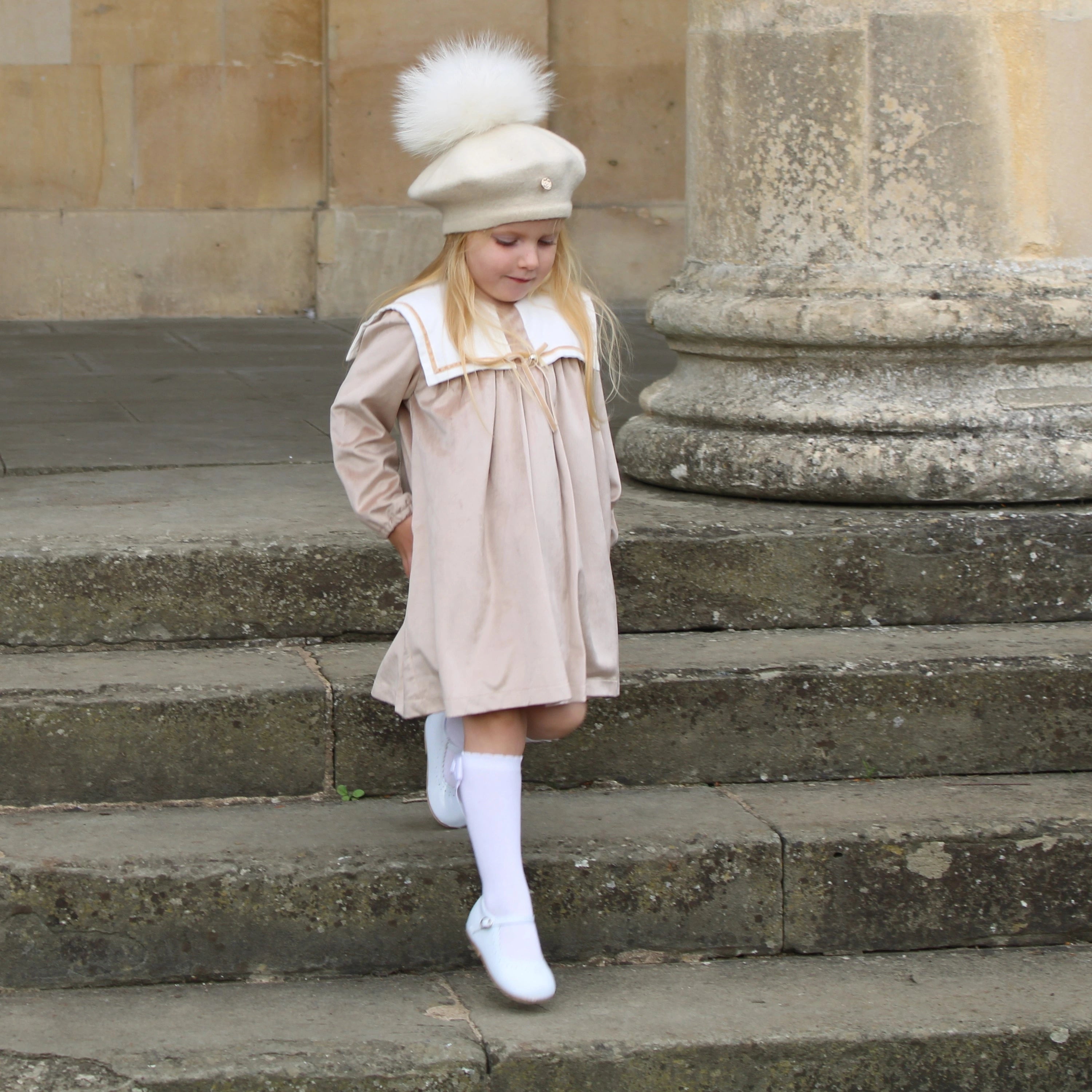 Young girl in a beige dress and white hat with a fur pom-pom standing on stone steps.