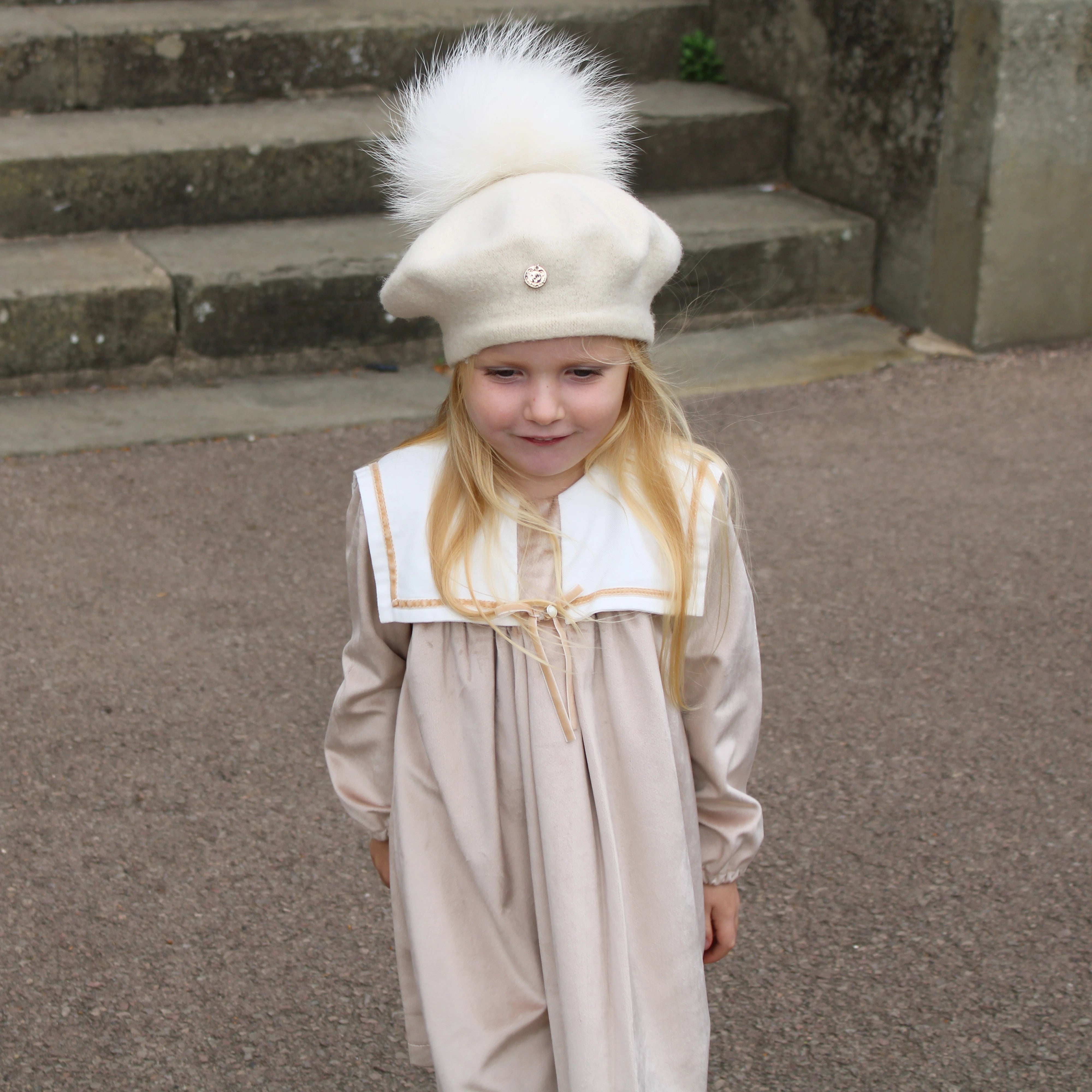 Young girl in a beige outfit with a white hat standing on a stone path.