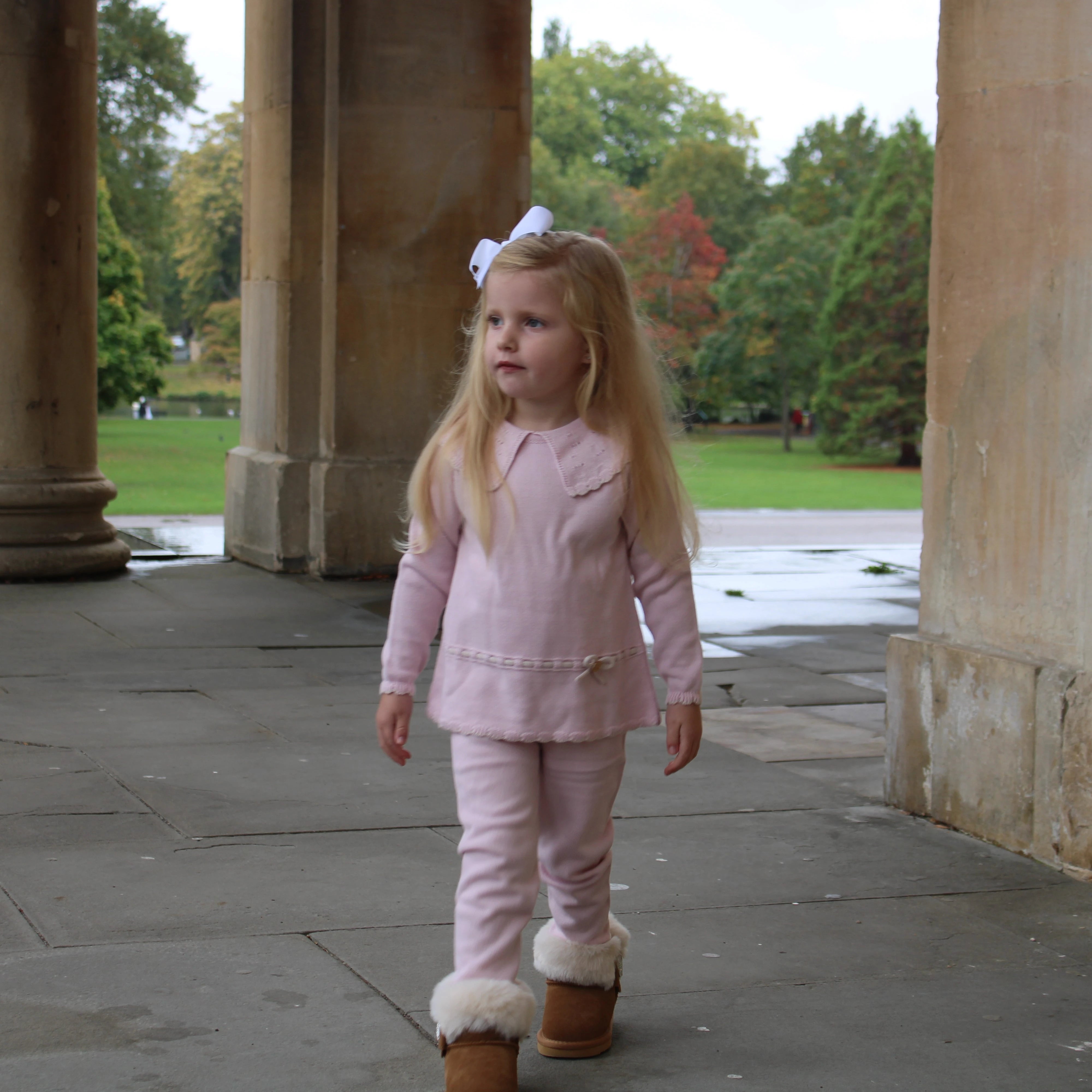 Young girl in pink outfit with fur-lined boots standing between stone columns.