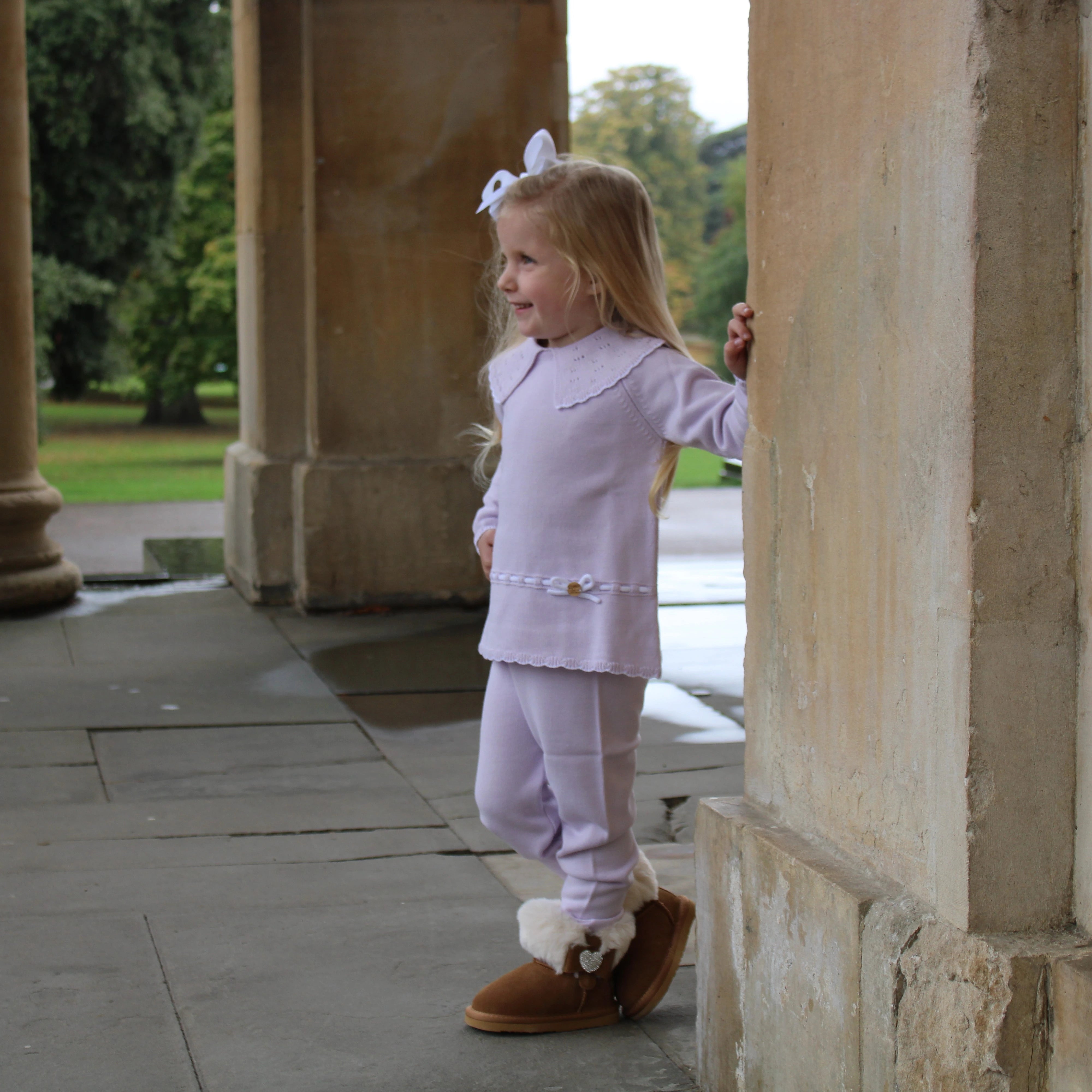 Child in a light purple outfit with fur-lined shoes standing on a stone path.