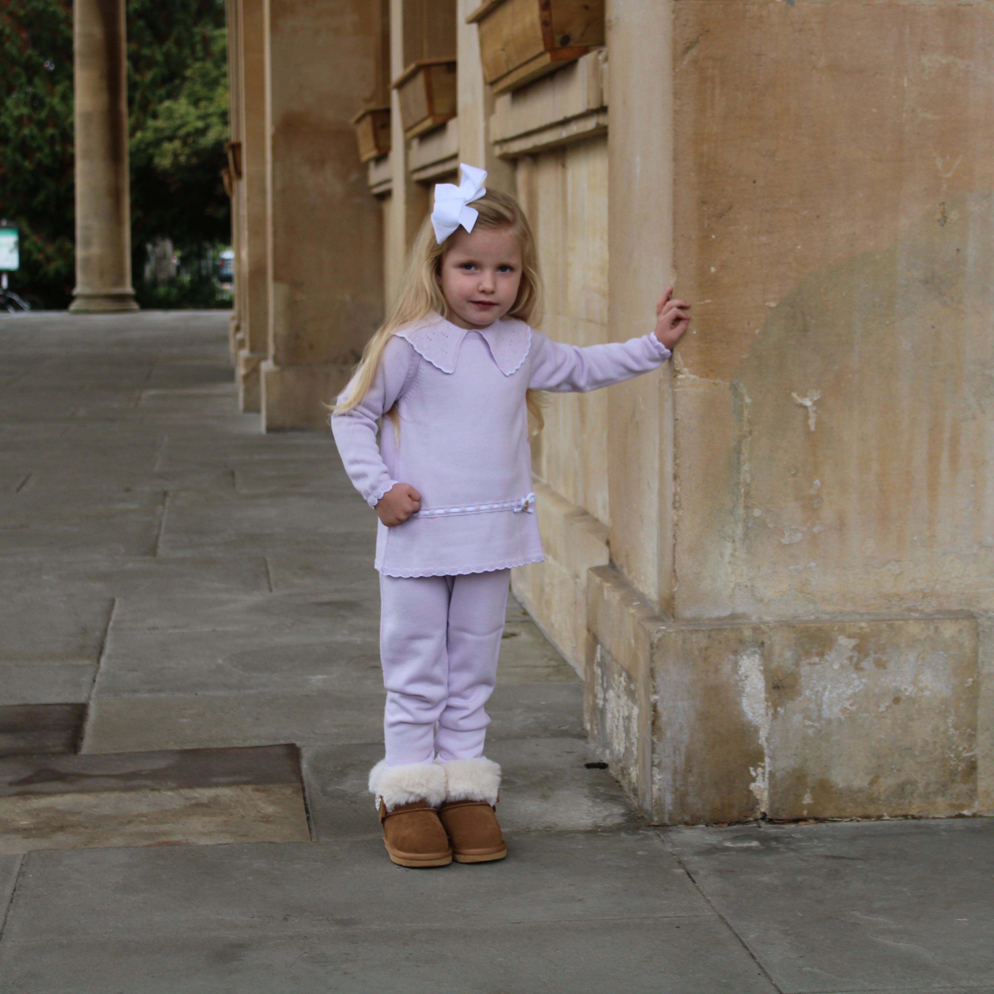 Young girl in a lavender outfit with brown boots standing against a stone wall.