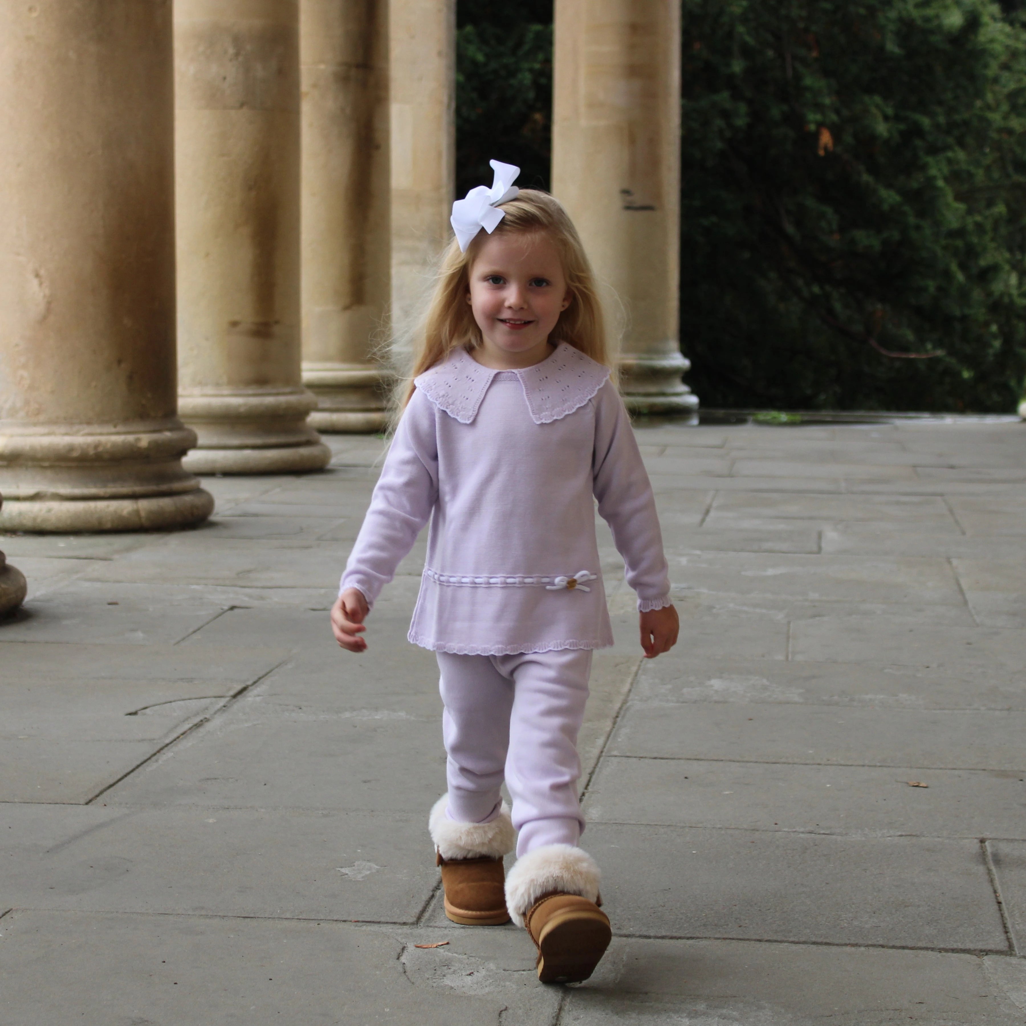 Young girl in a light purple outfit with white collar and brown boots walking on a stone path.