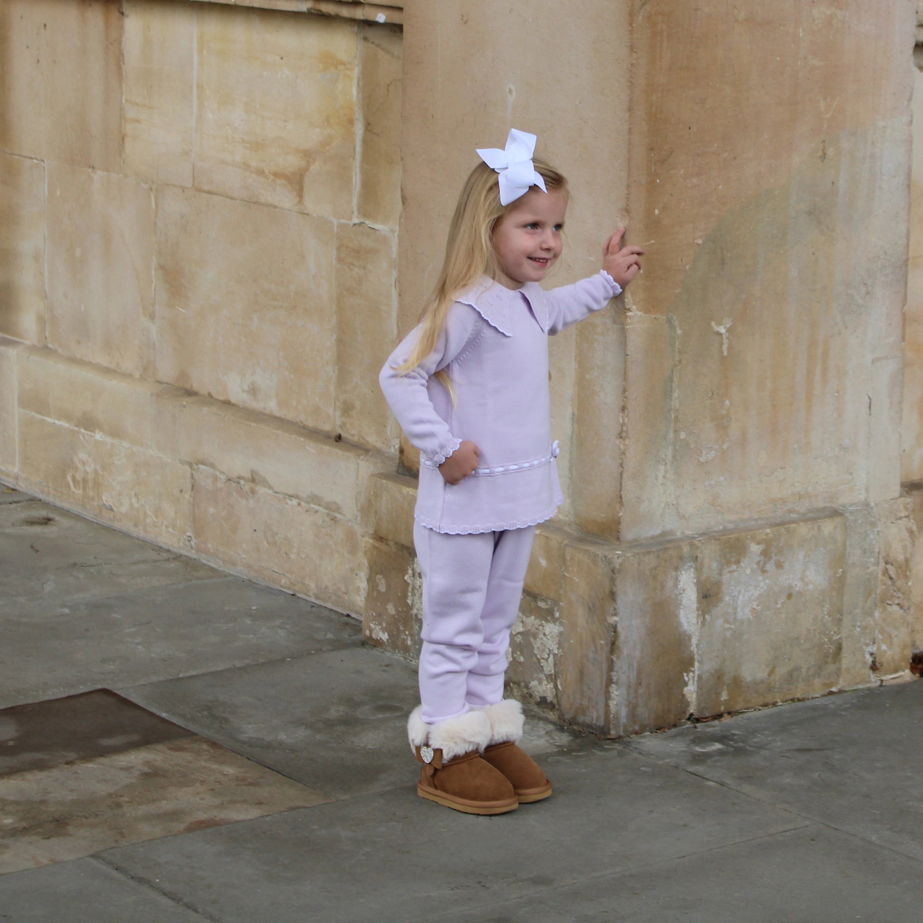 Young girl in a light purple outfit with white fur trim and brown boots standing against a stone wall.