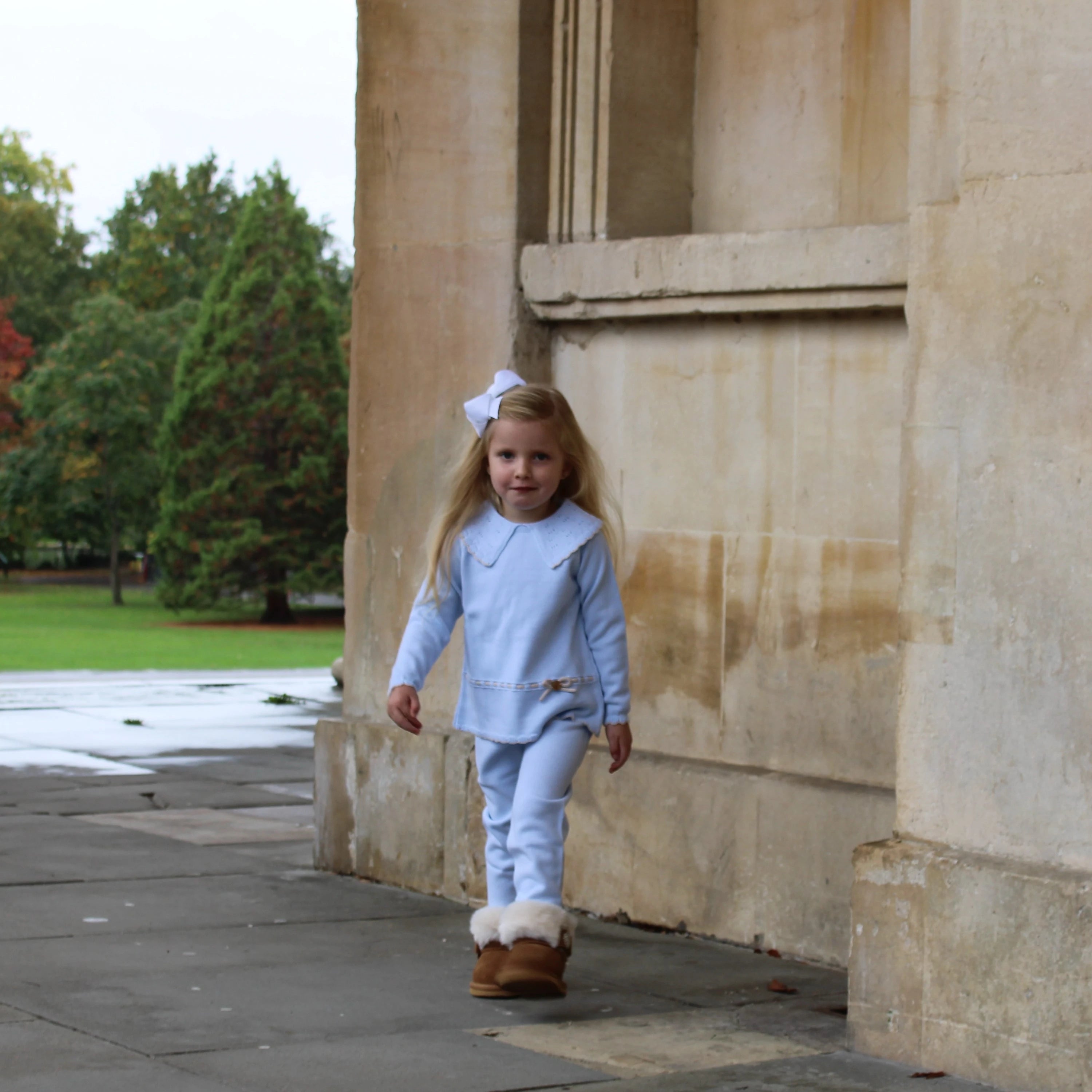 Young girl in a light blue outfit with white fur trim walking near stone columns.