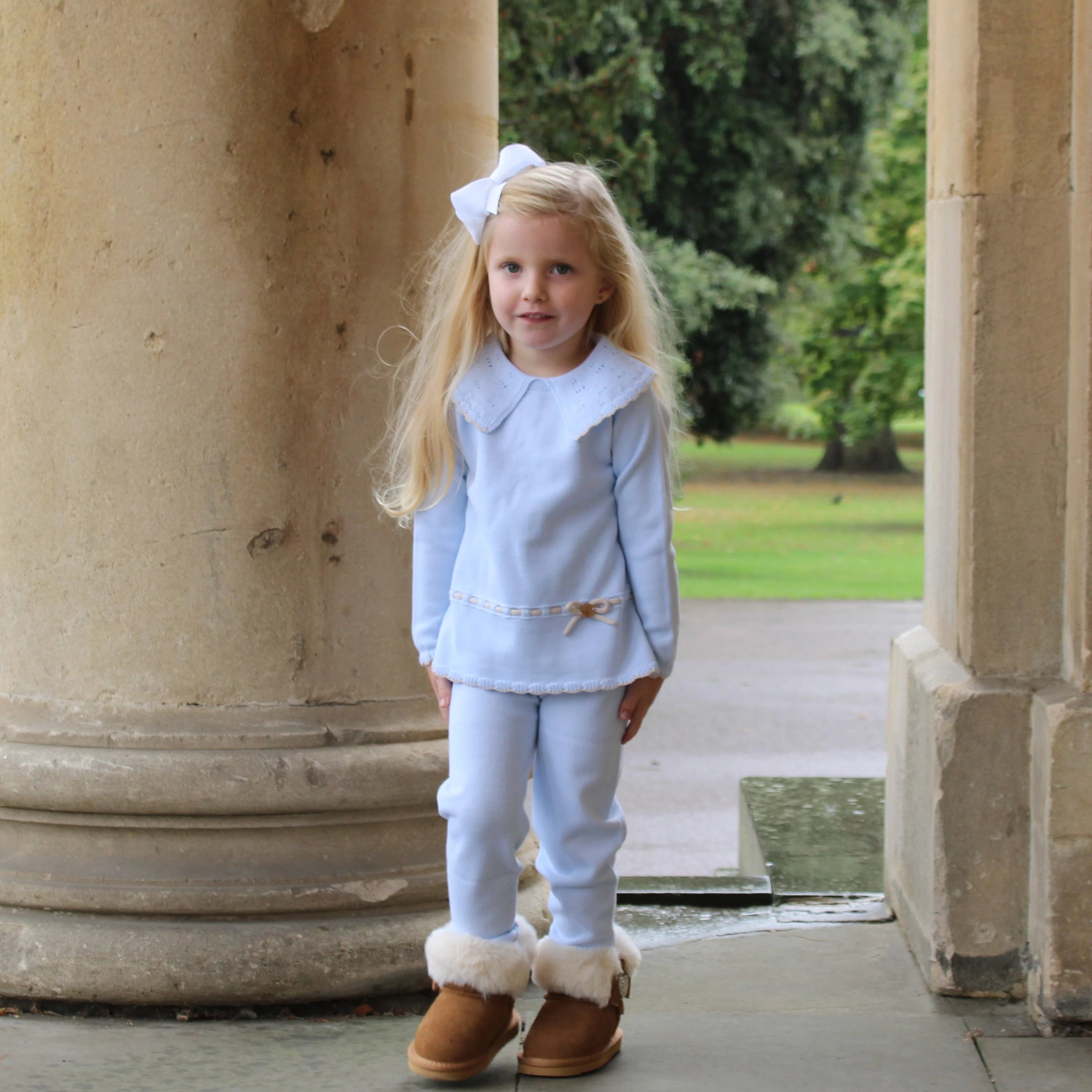 Young girl in a light blue outfit with brown boots standing in front of stone columns.