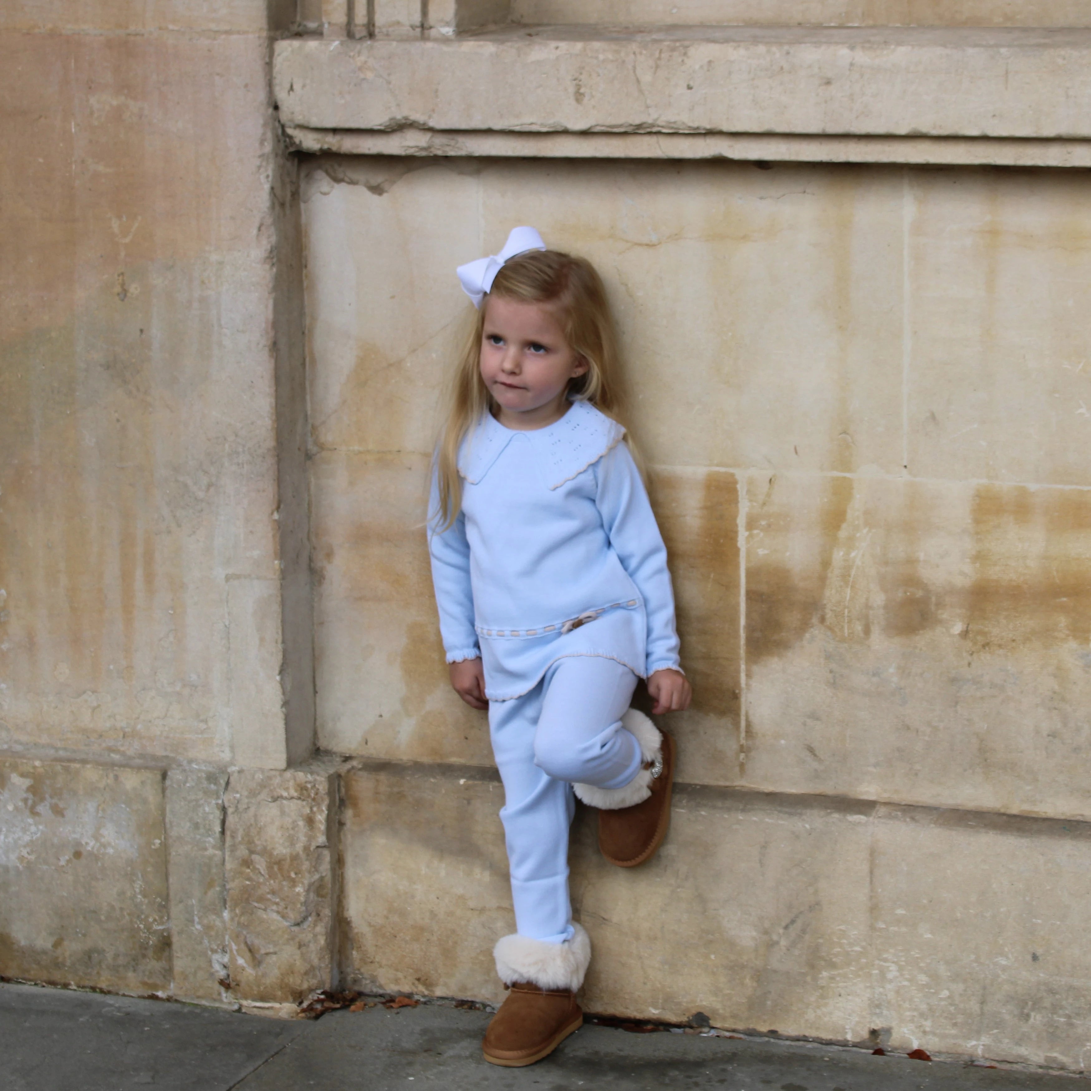 Young girl in a light blue outfit with brown boots standing in front of stone columns.