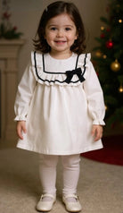 Child wearing a white dress with black trim and a bow in front of a Christmas tree.