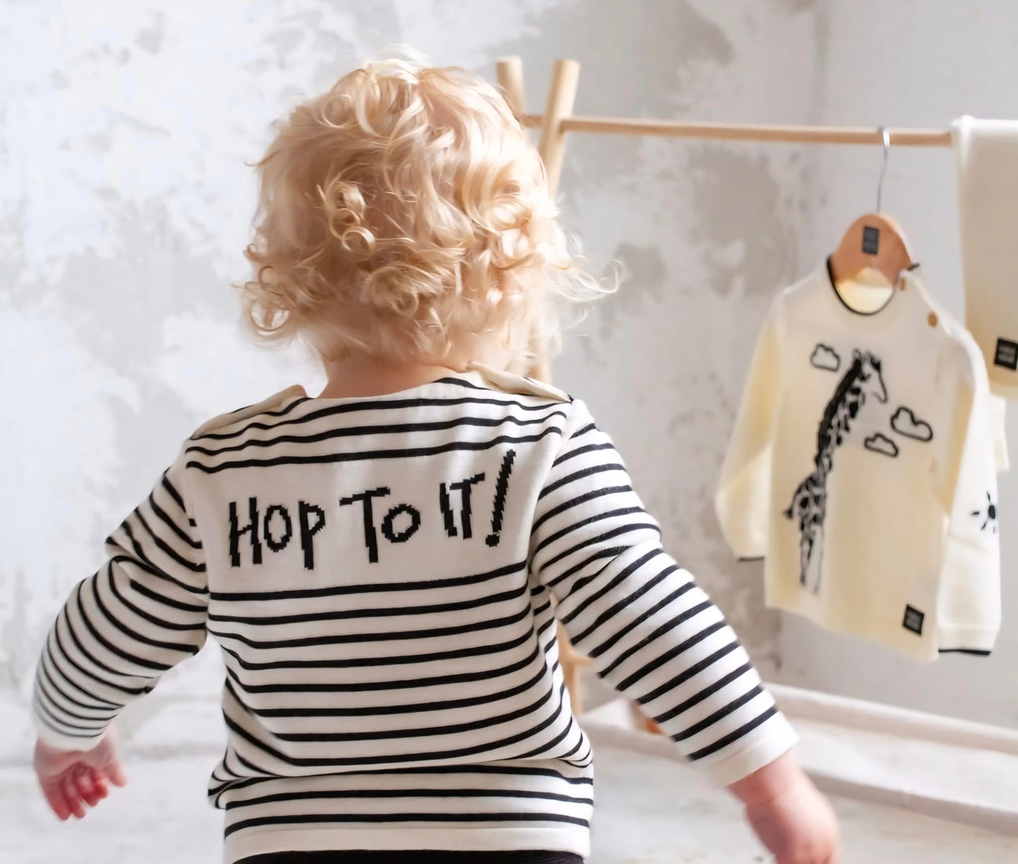 Child wearing a striped shirt with 'Hop to it!' text, standing in front of a clothing rack with baby clothes.