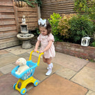Child in a garden pushing a toy shopping cart with a teddy bear inside.