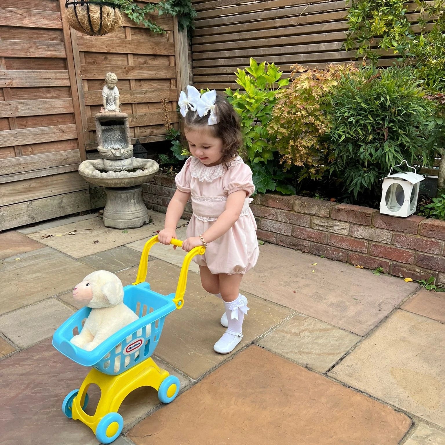 Child in a garden pushing a toy shopping cart with a teddy bear inside.