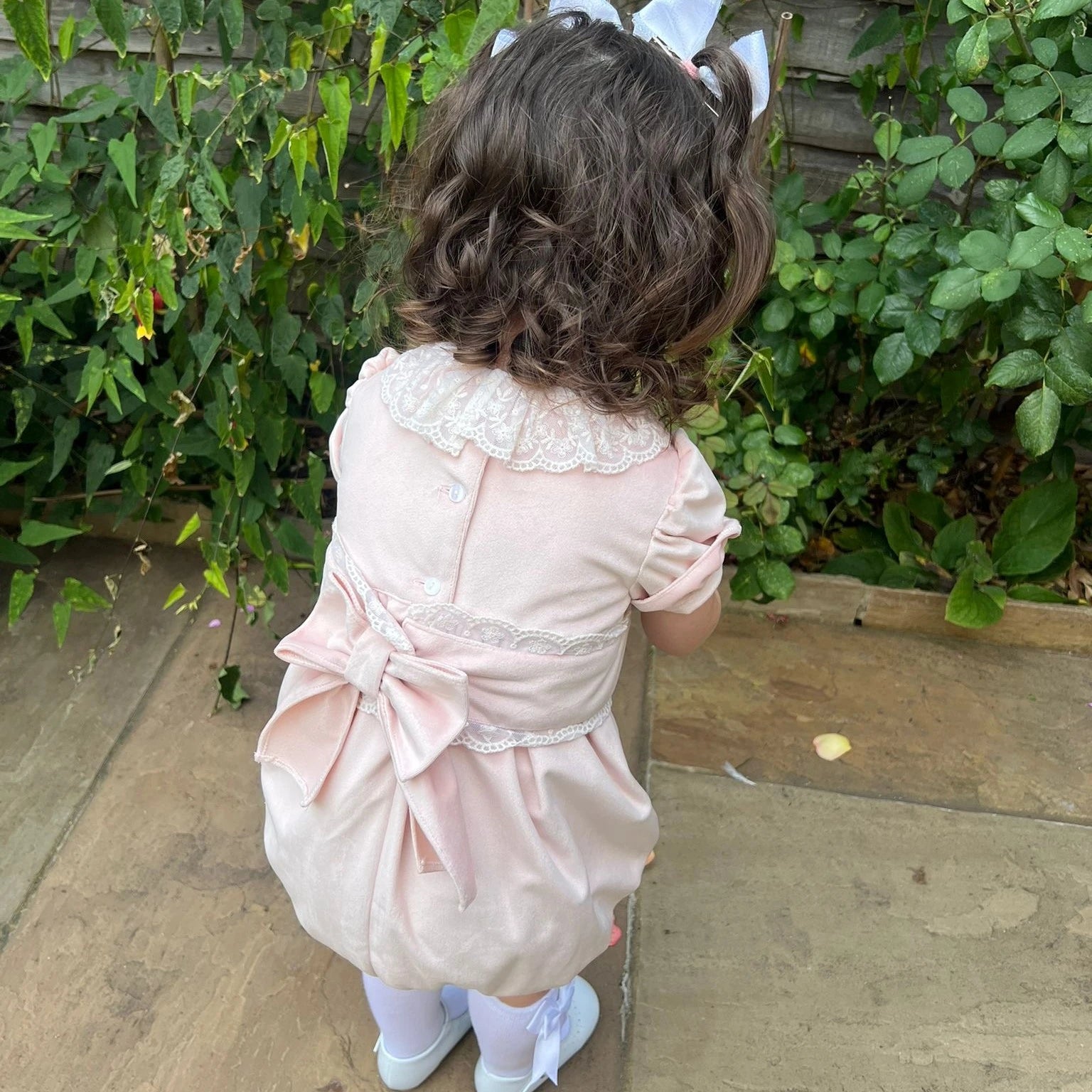 Child in a pink dress with a large bow standing outdoors on a patio.