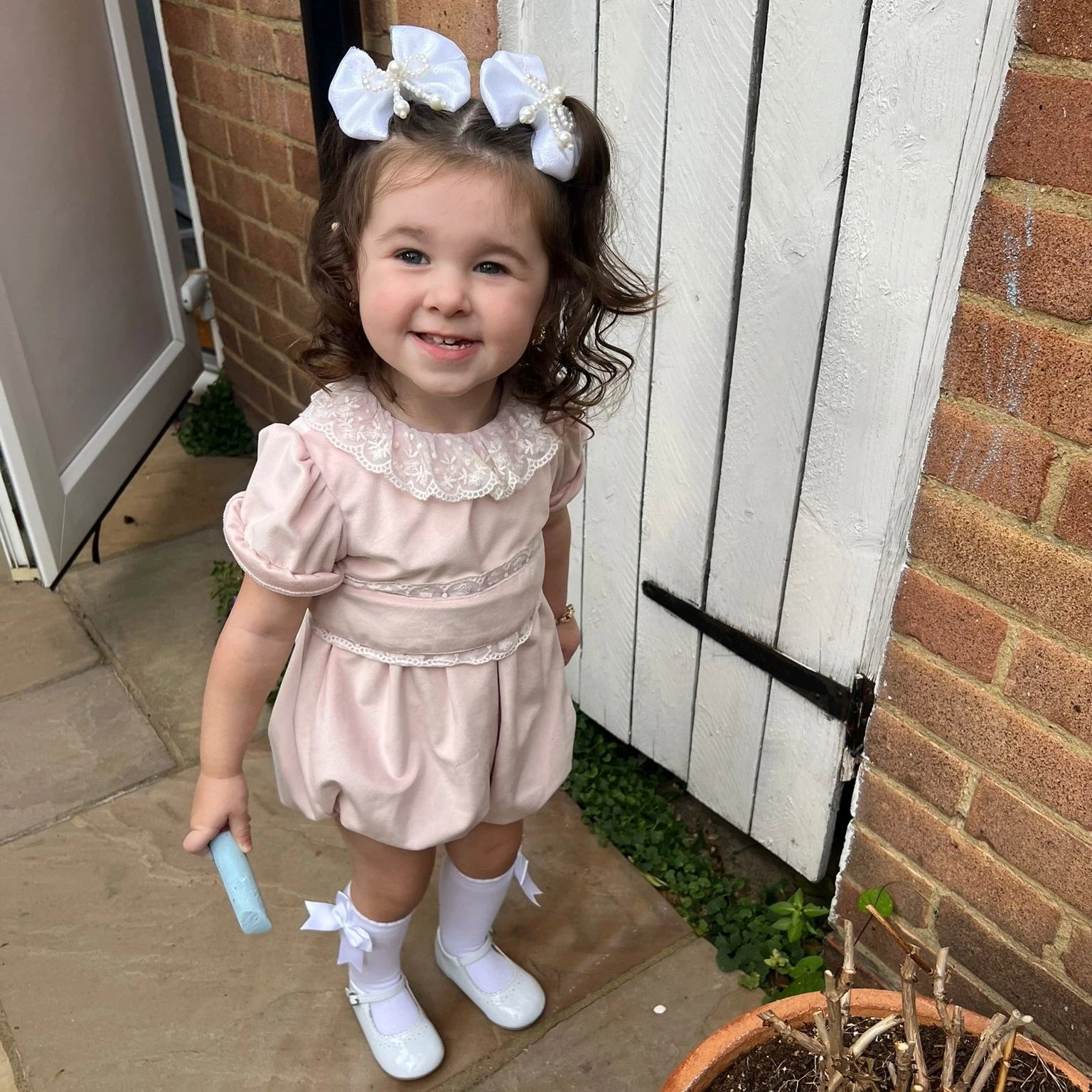 Young girl in a pink dress with white bows standing outside a house.