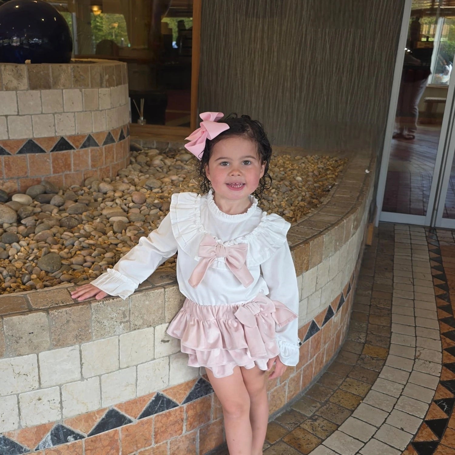 Young girl in a white and pink outfit with bows standing near a decorative stone wall.