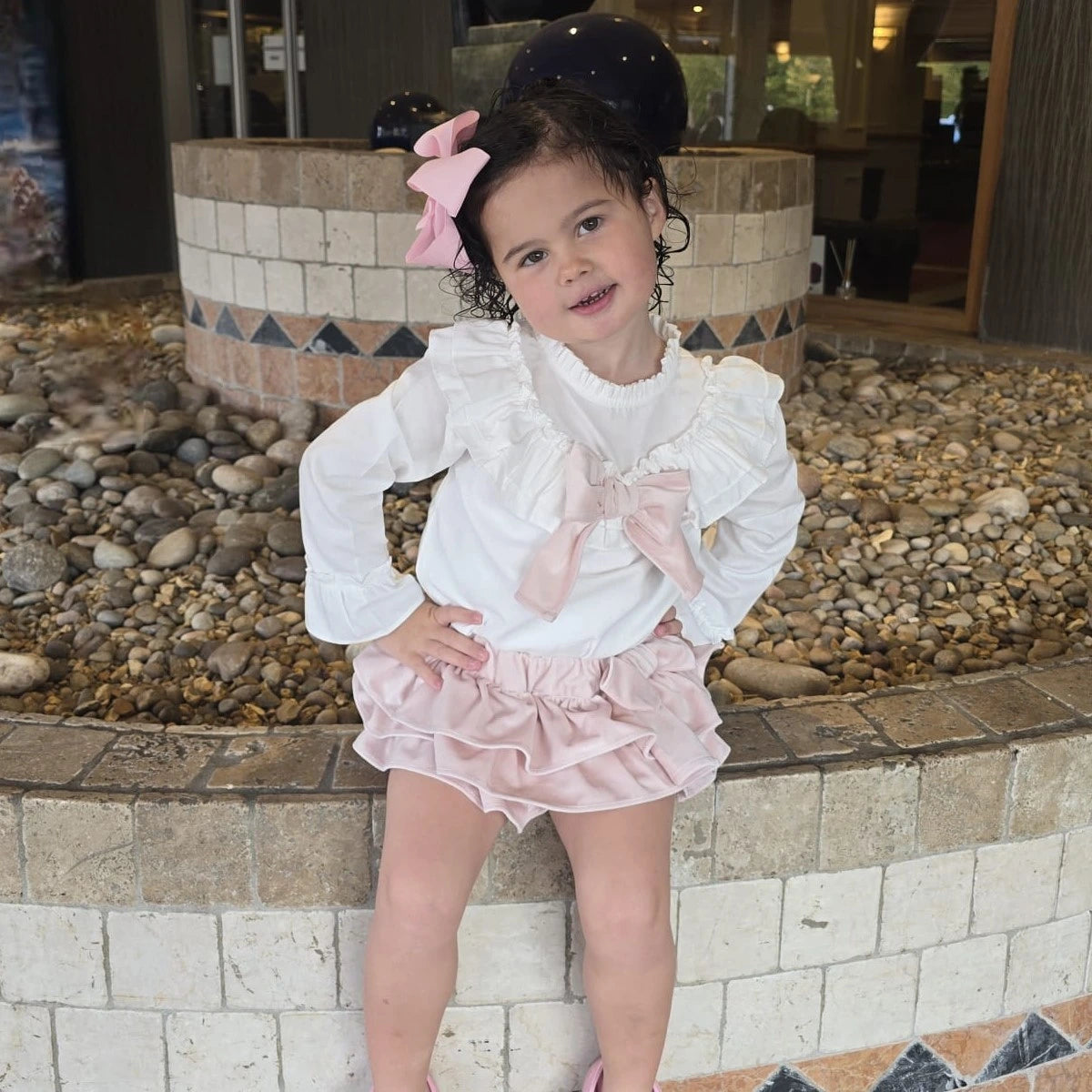 Young girl in a white blouse and pink skirt standing in front of a decorative fountain.