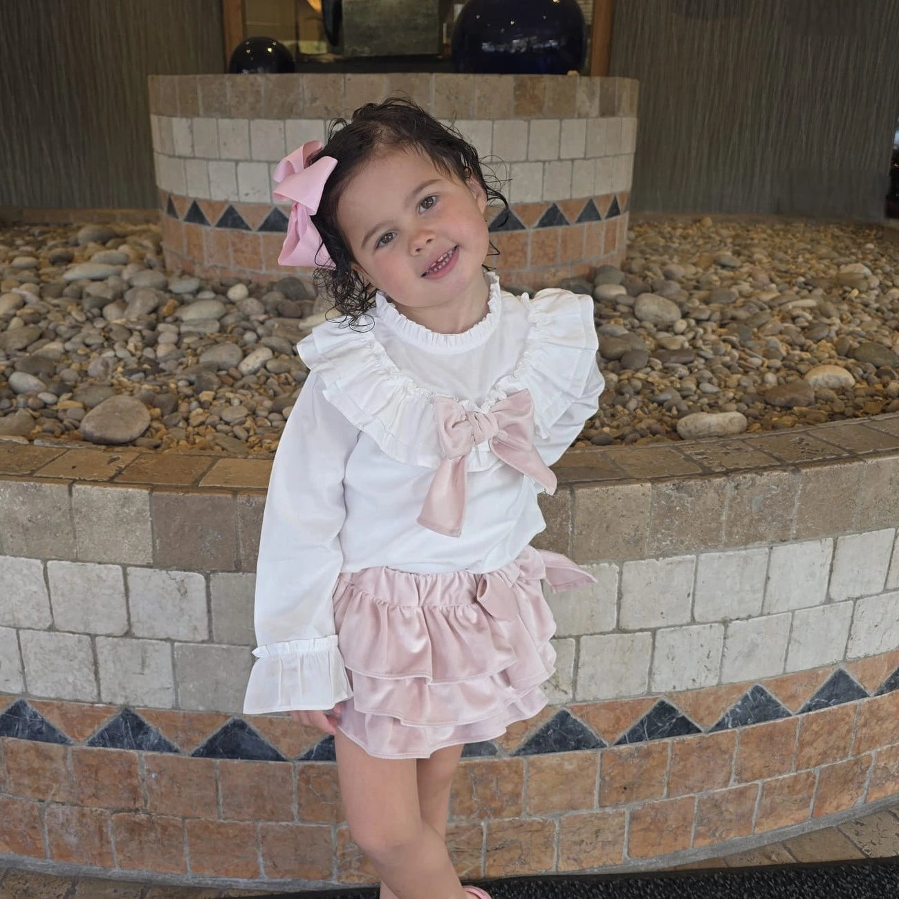 Young girl in a white blouse with ruffles and pink skirt standing in front of a stone wall.