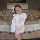 Young girl in a white blouse with ruffles and pink skirt standing in front of a stone wall.